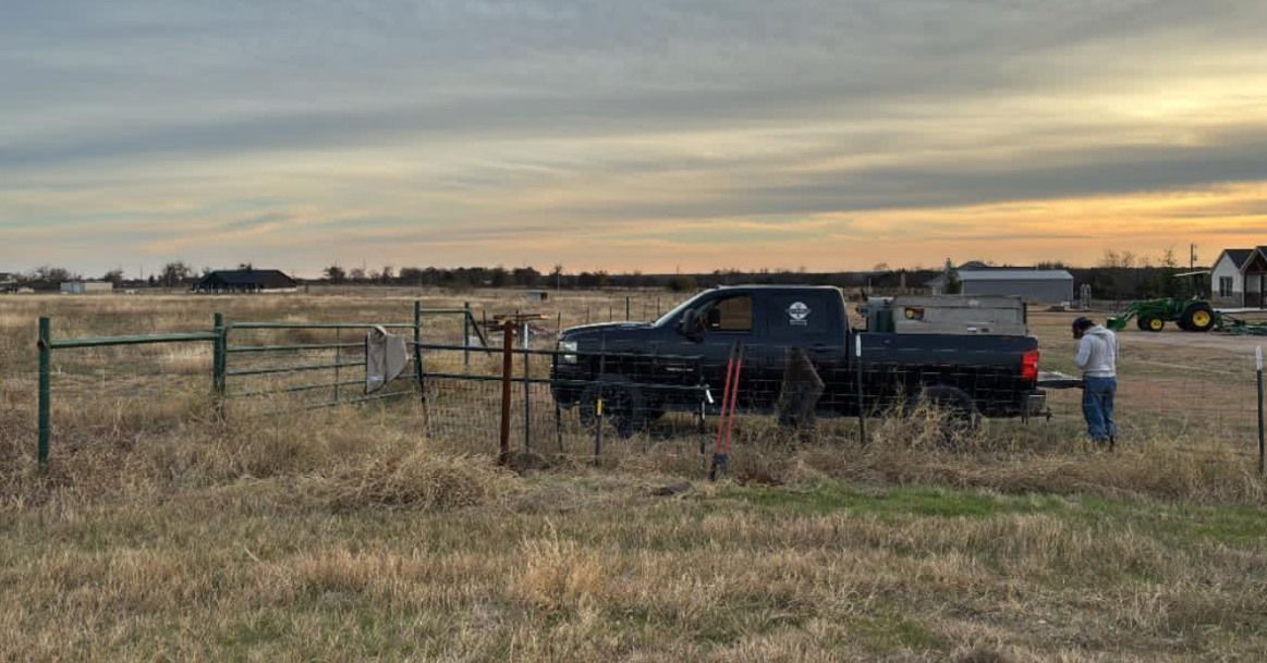 Black pickup truck in a fenced field at sunset, with two people standing nearby.