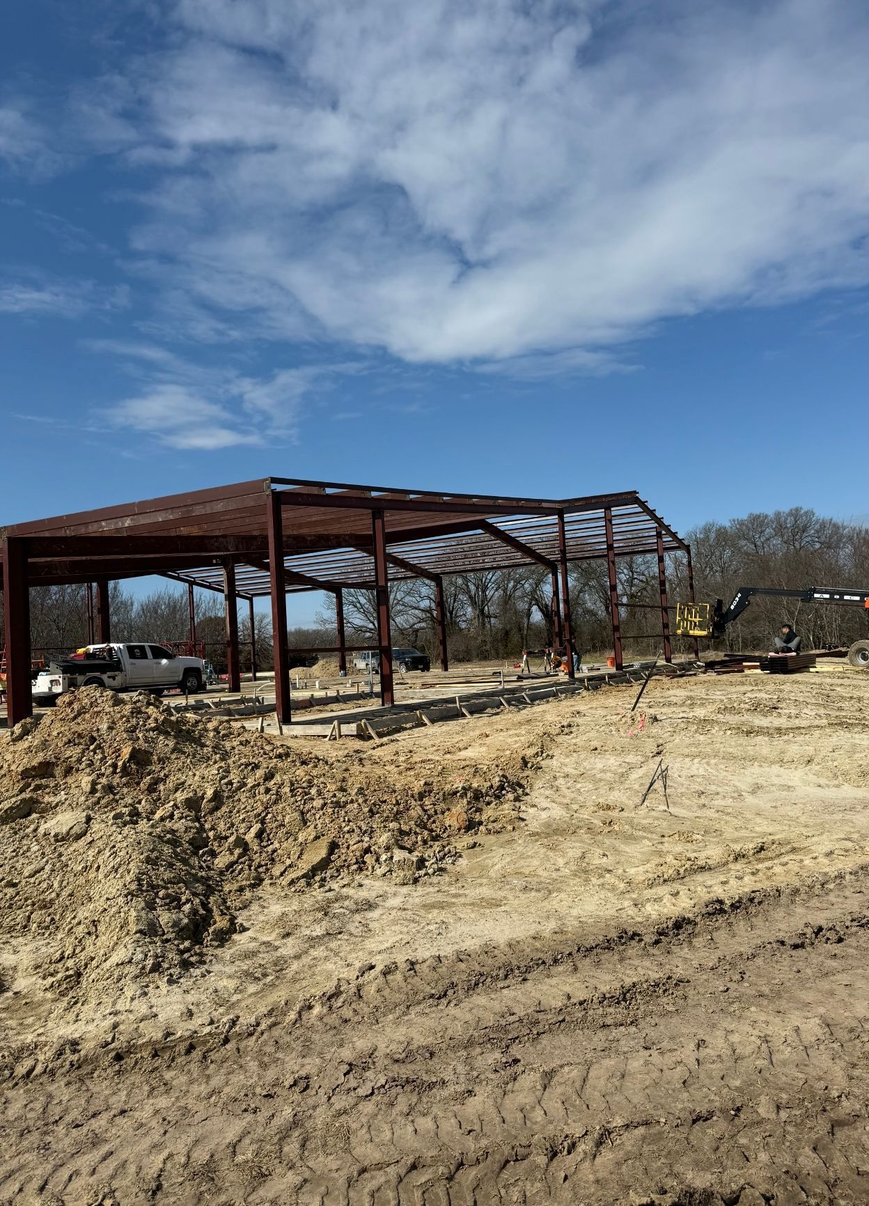 Rust-colored steel frame building under construction on a dirt lot beneath a cloudy blue sky