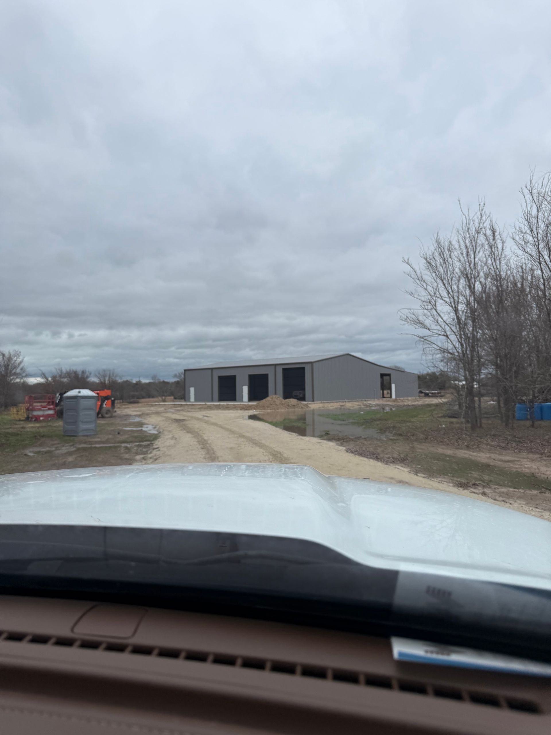 Driveway view of a gray barn-like building under a cloudy sky, seen over a vehicle hood.