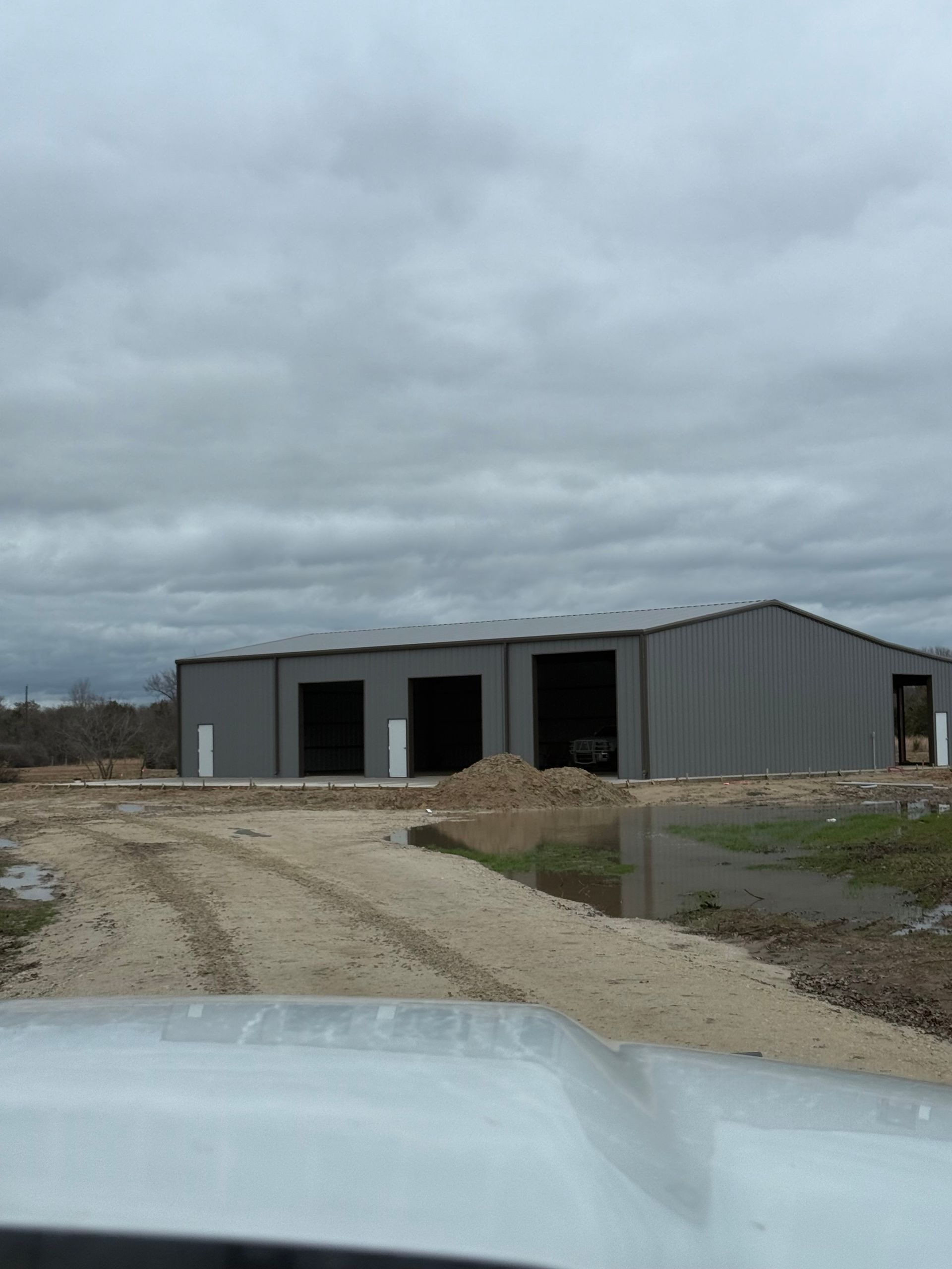 Gravel driveway leading to a gray metal warehouse under a cloudy sky, with a car hood in the foreground