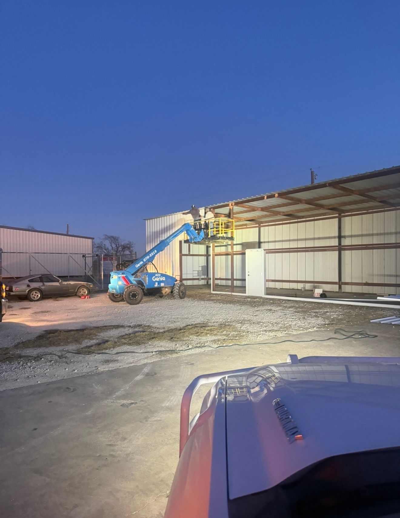 Blue lift beside a warehouse at dusk, with a truck hood in the foreground and gravel lot around it