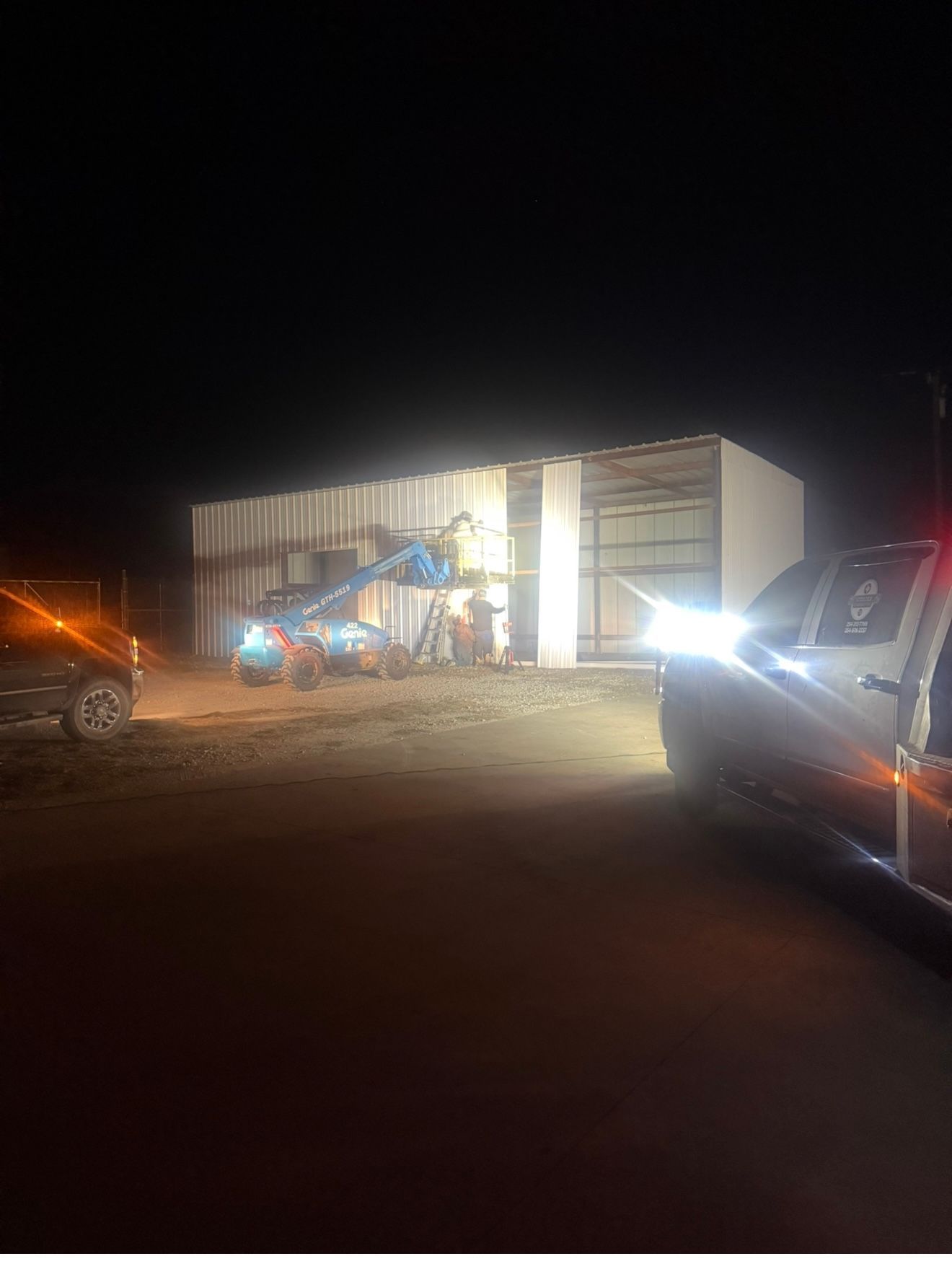 Nighttime construction scene with a lit building, excavator, and parked vehicles on a dirt lot.