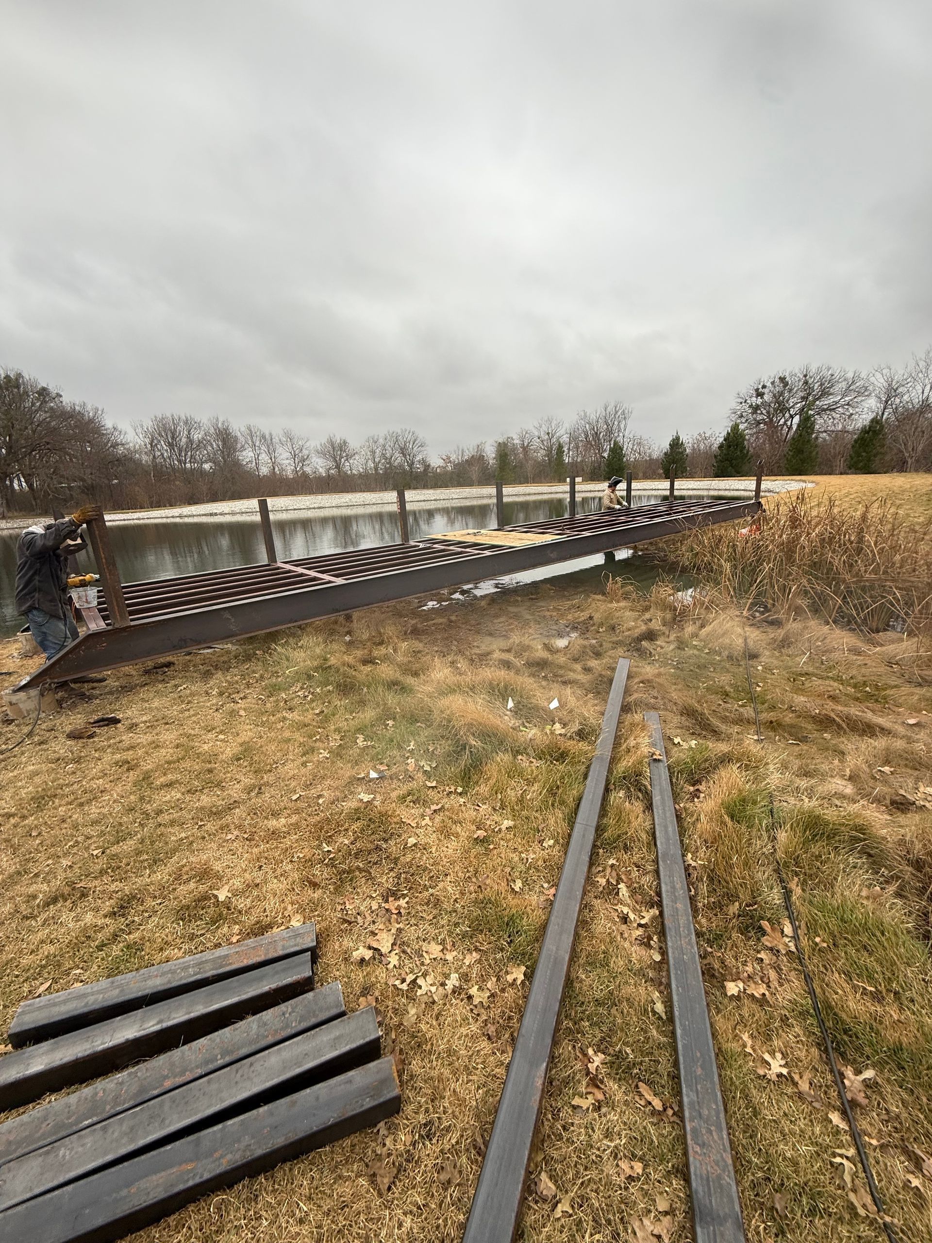 Damaged riverside fence along a grassy path under a gray sky, with a river in the background