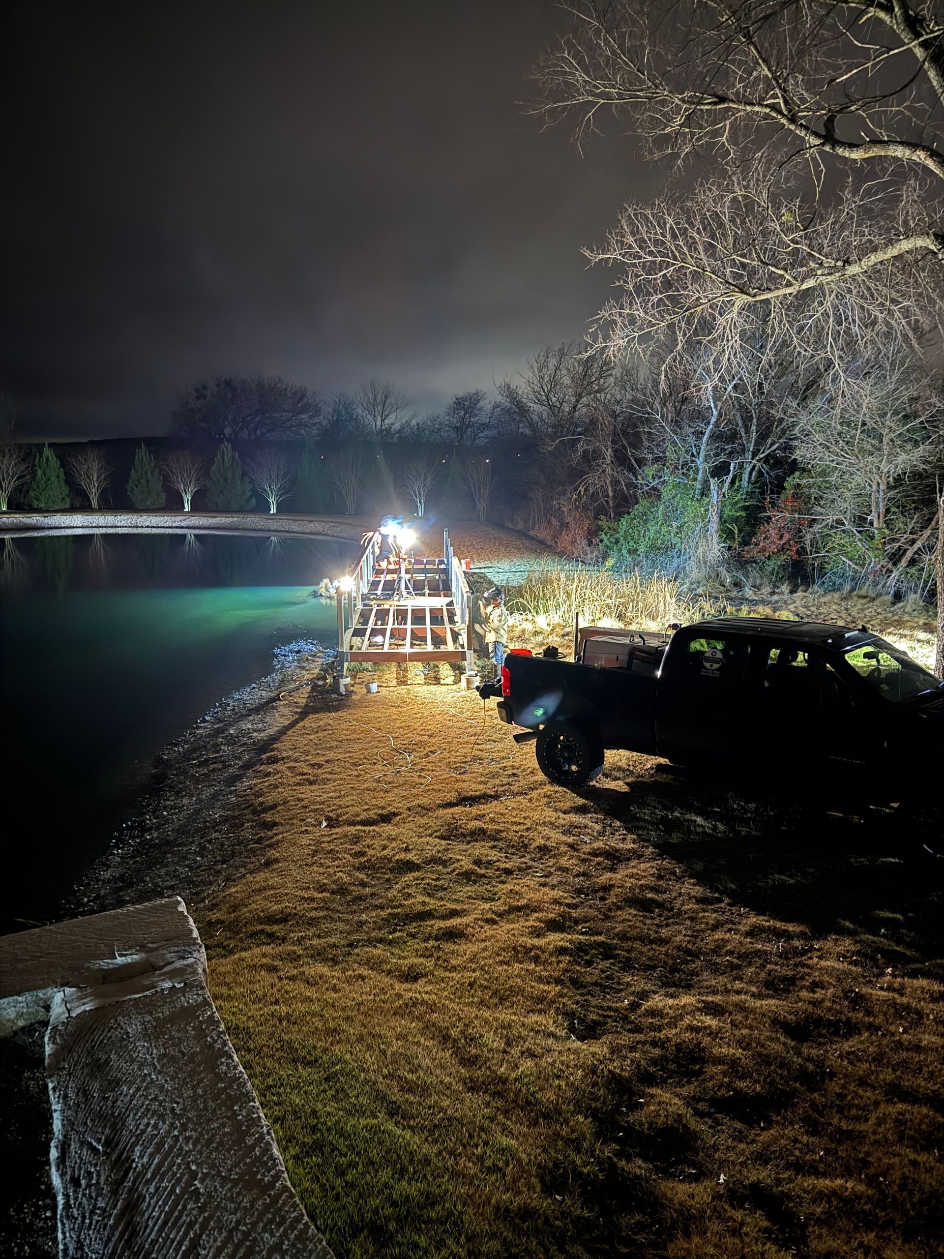 Nighttime riverside scene with a lit dock, parked vehicle, and bare trees reflected on calm water