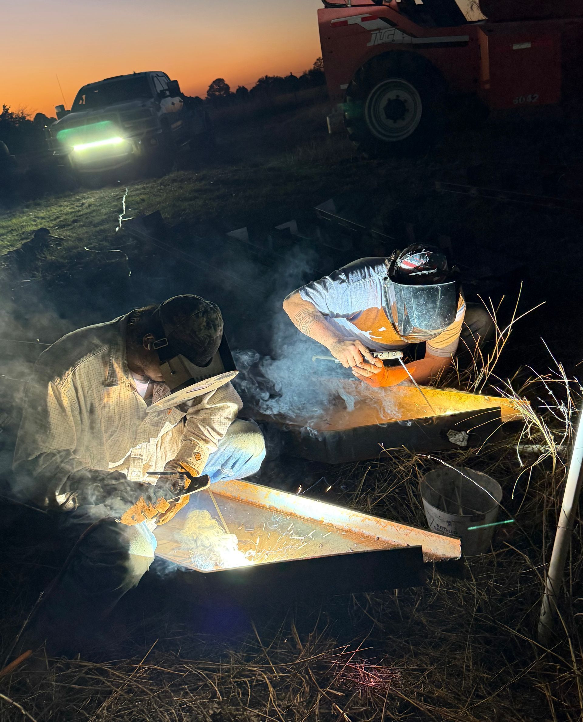 Person in reflective gear works by firelight near smoking debris at dusk, with a vehicle in the background