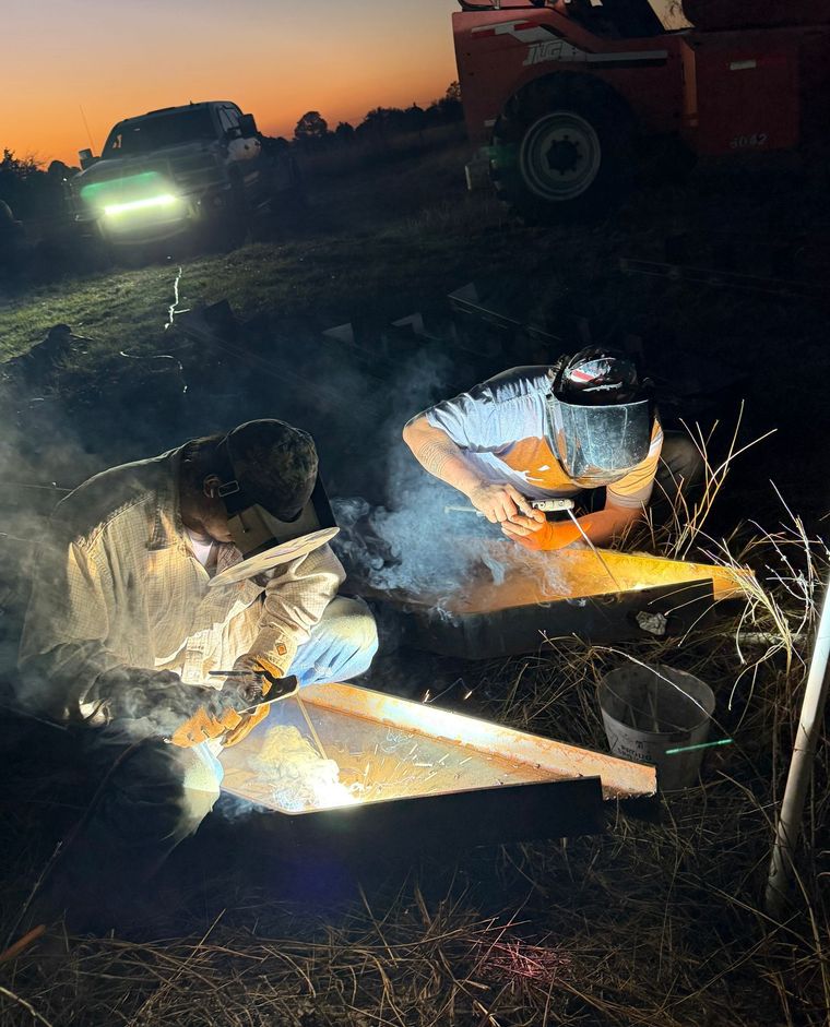 Worker in reflective gear kneeling by a smoky fire at dusk, with machinery and a vehicle in the background