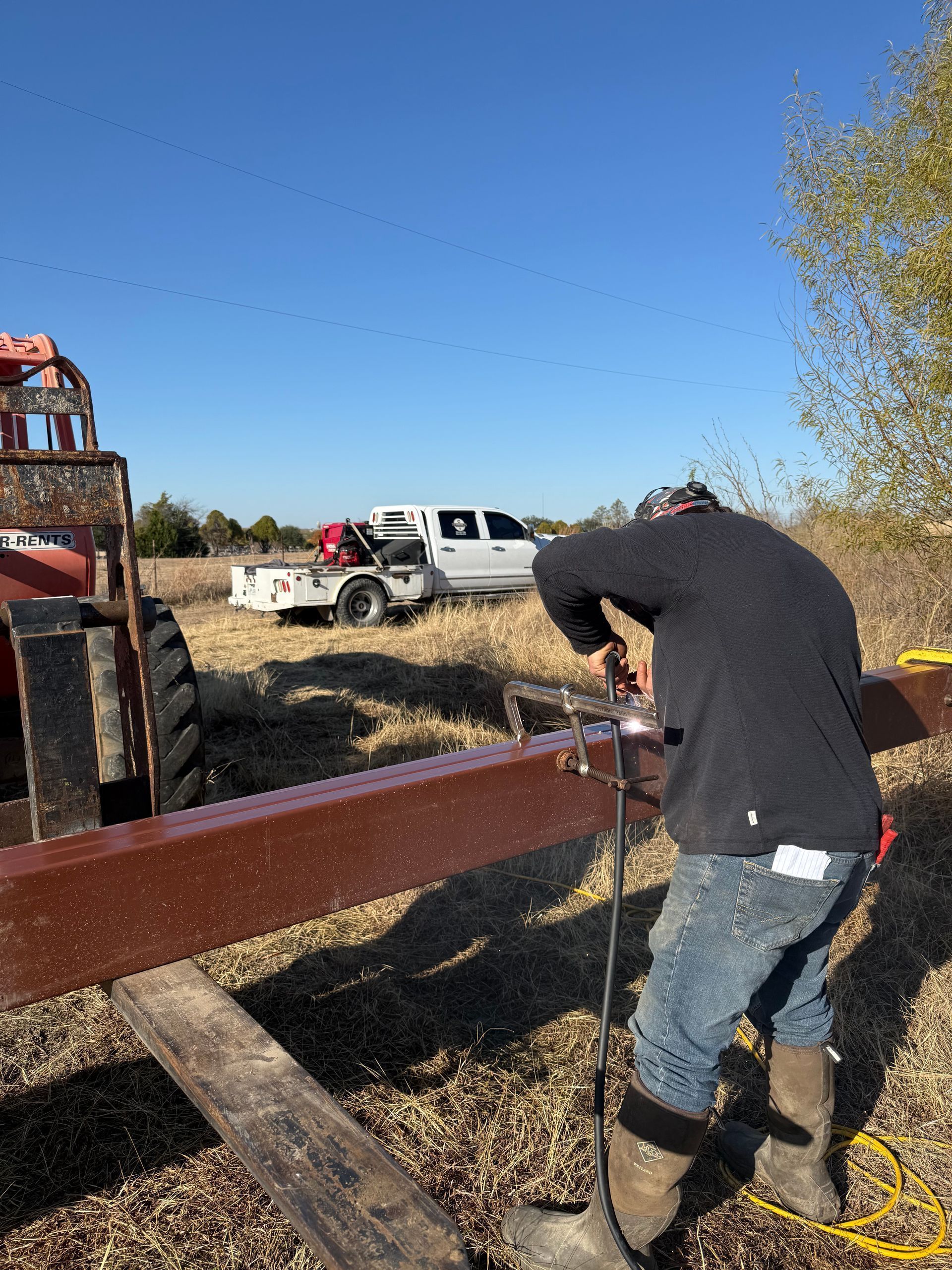 Person drilling into a red steel beam at a rural worksite with a white truck and equipment in the background