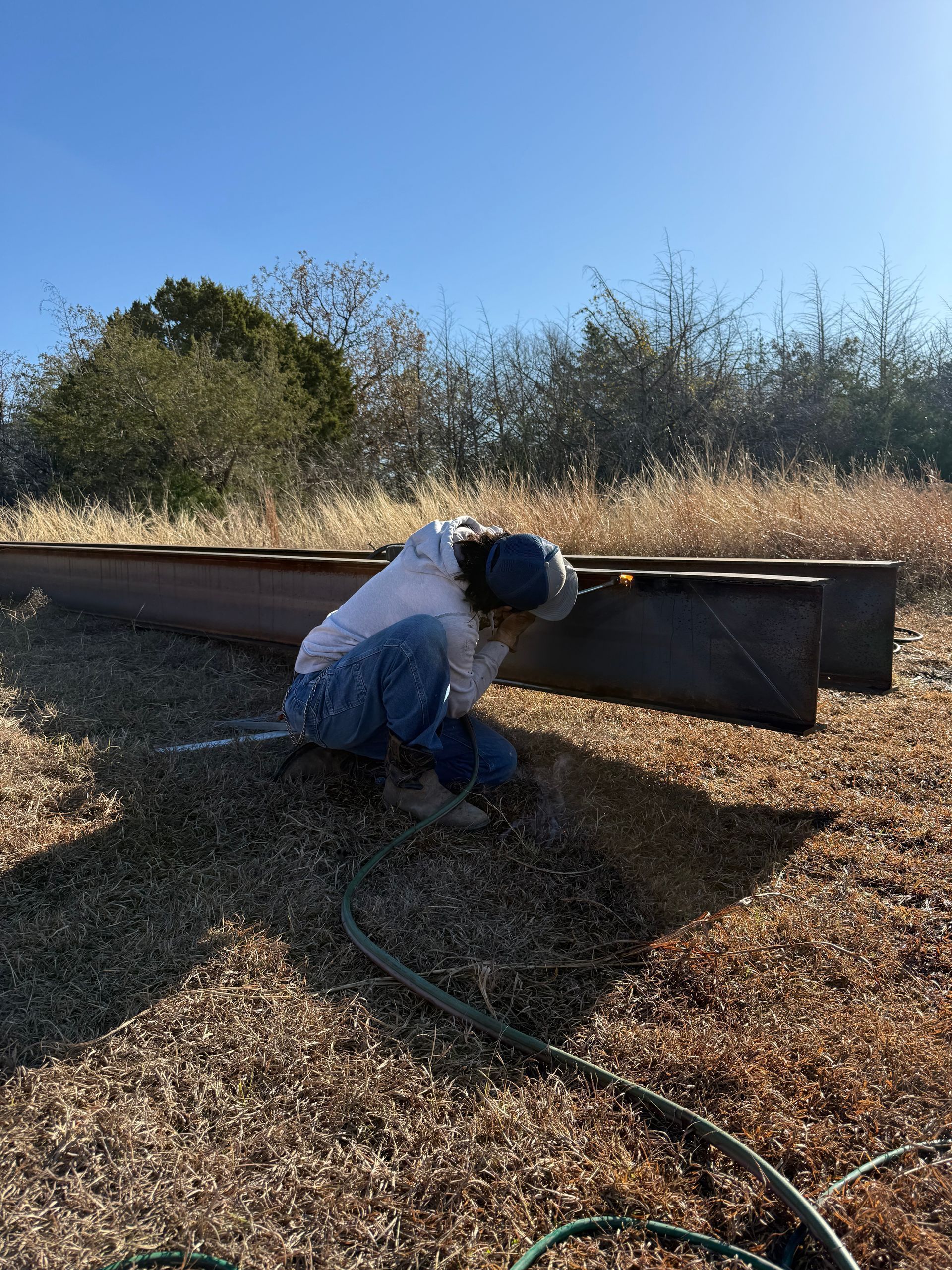 Person kneeling beside a raised garden bed, working in a sunny outdoor yard with hose on wood chips