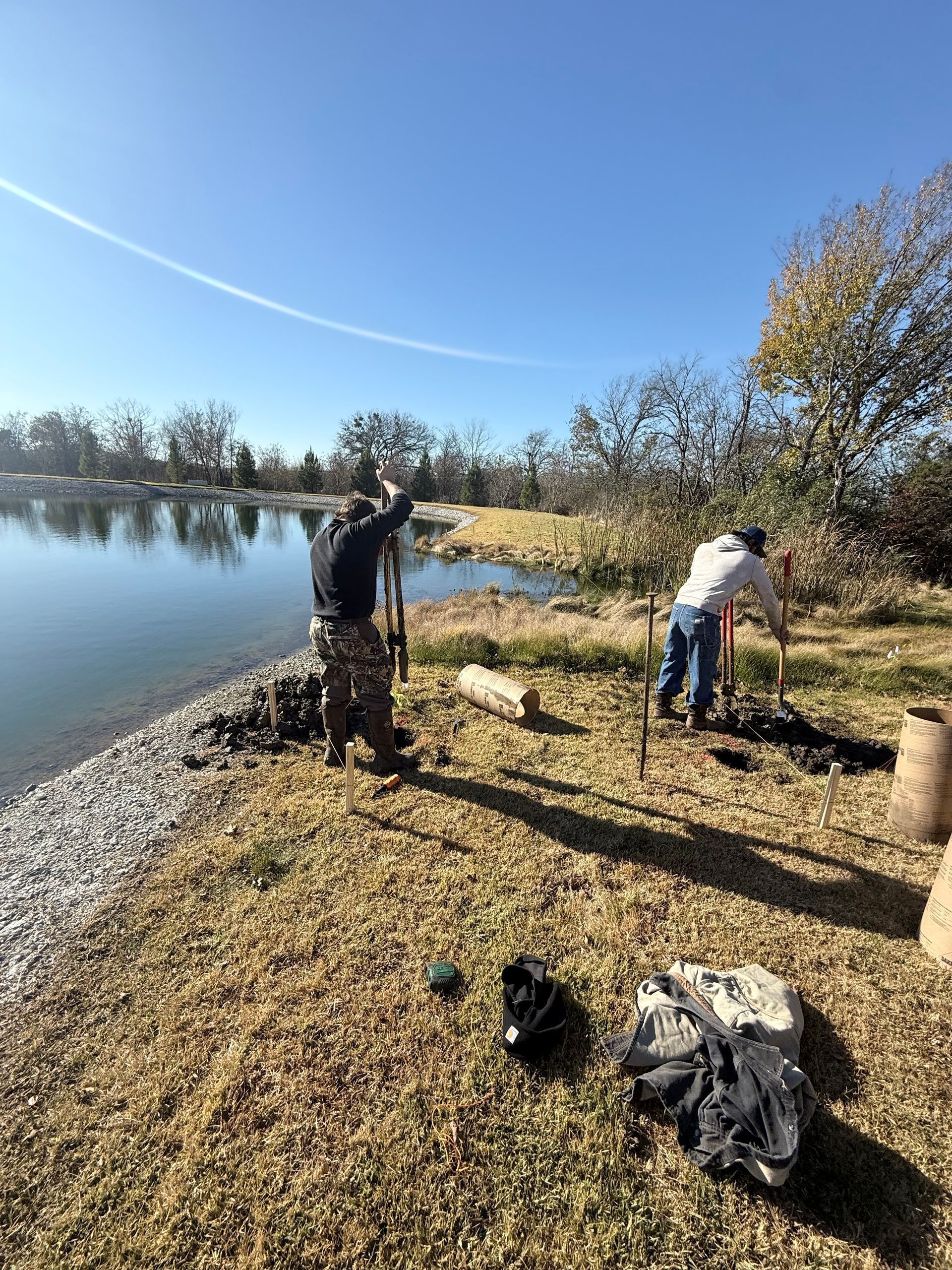 Two people working by a lakeshore on a sunny day, with gear on the ground and trees in the background.