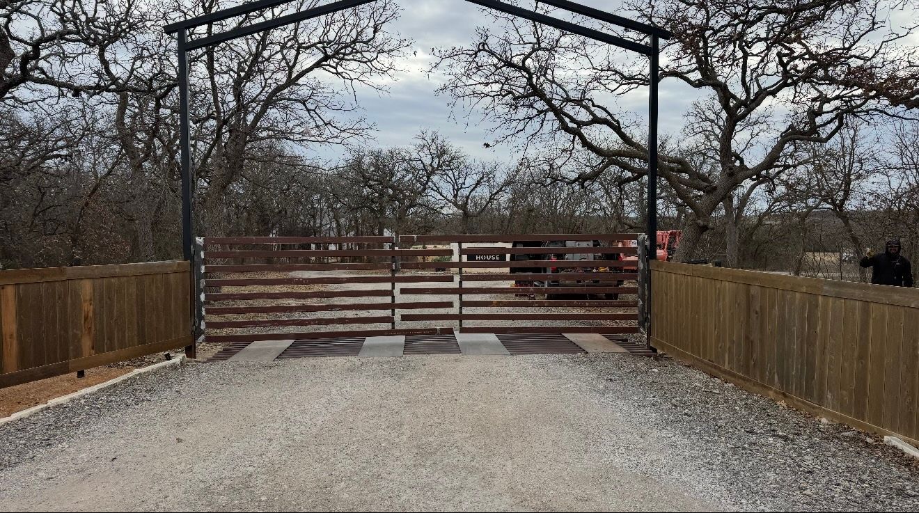 Gravel driveway leading to a wooden gate and fence under leafless trees, with a metal arch overhead
