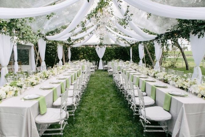 An outdoor reception tent with two long, white-clothed tables, green napkins, white chairs, and greenery hanging overhead.