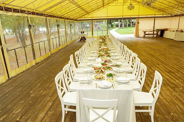 Long white table set for a meal inside a yellow tent with clear side panels, wooden floor, and a park view.