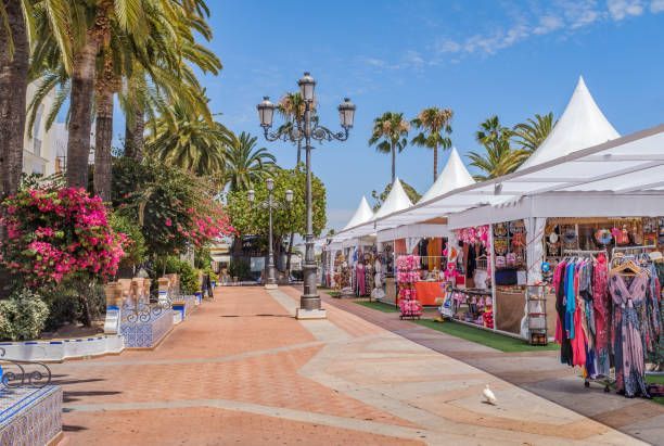 Outdoor market with white tents, vendors, and shoppers on a sunny walkway lined with palm trees and flowers.