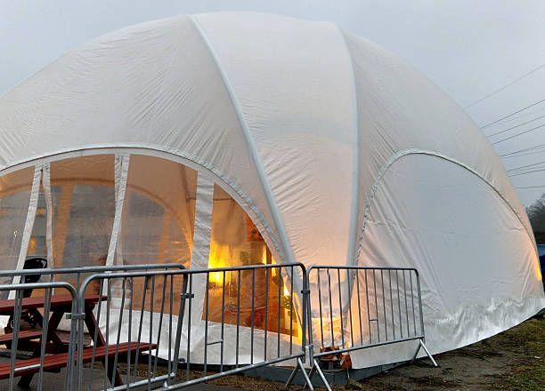 White, arched tent with transparent windows and interior lighting, next to a metal barrier and outdoor picnic table.