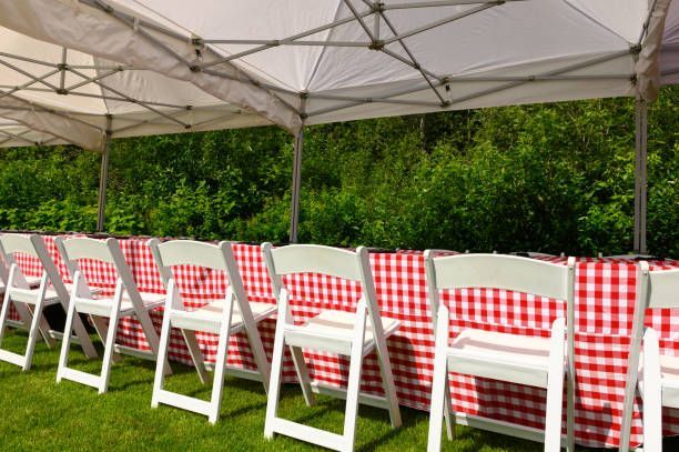 White chairs line a table with red gingham cloth under a white tent on grass, with green bushes in the background.