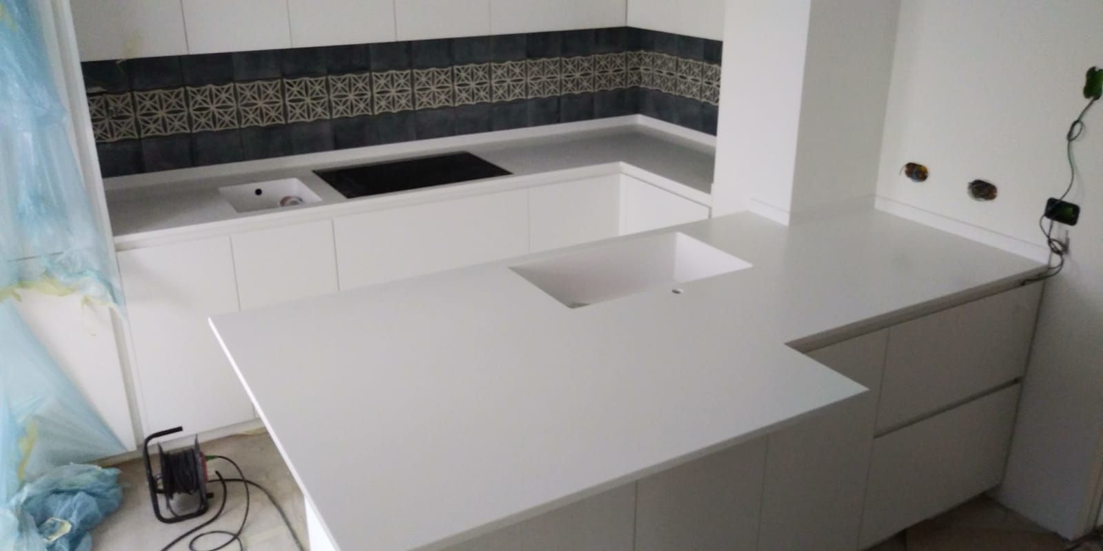 Una moderna cucina bianca con piano di lavoro e paraschizzi in piastrelle scure; cavi e utensili visibili.