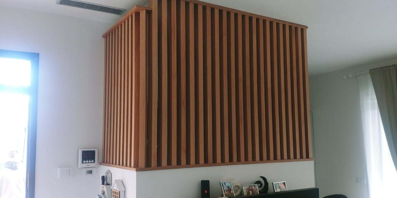 Dettaglio architettonico con doghe in legno, probabilmente un assorbitore acustico, su un pilastro bianco in una stanza con finestra e tende.