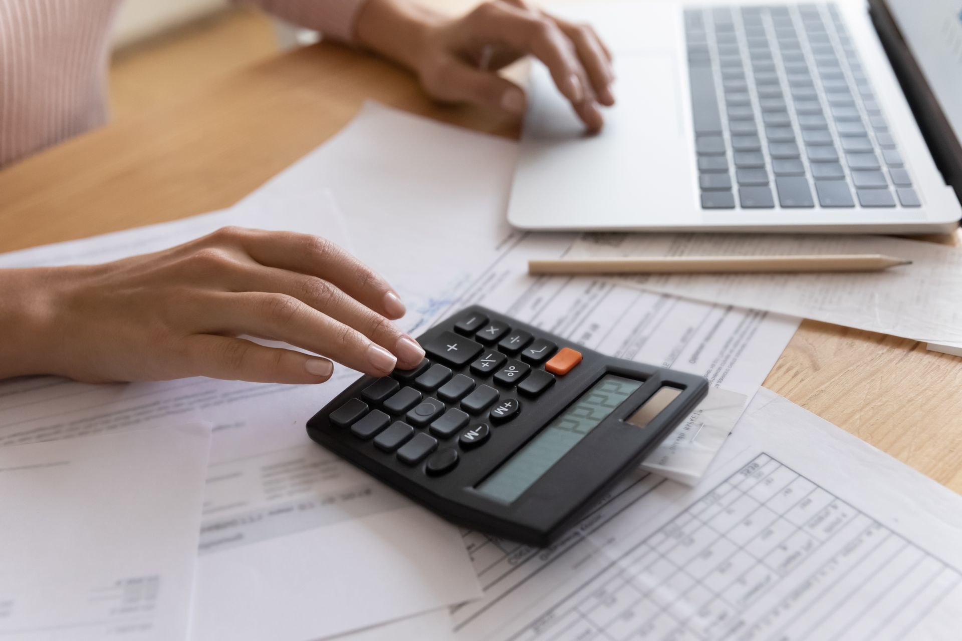 Hands using a calculator beside a laptop on paperwork at a desk