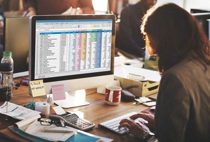 Person working at a desk with a large monitor showing a spreadsheet in a busy office