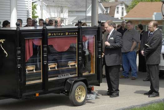 A man in a suit stands in front of a black coffin on a trailer.