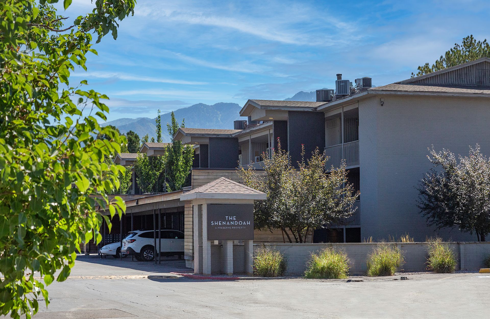 Apartment complex with a sign, parking, trees, and mountains in the background under a blue sky.