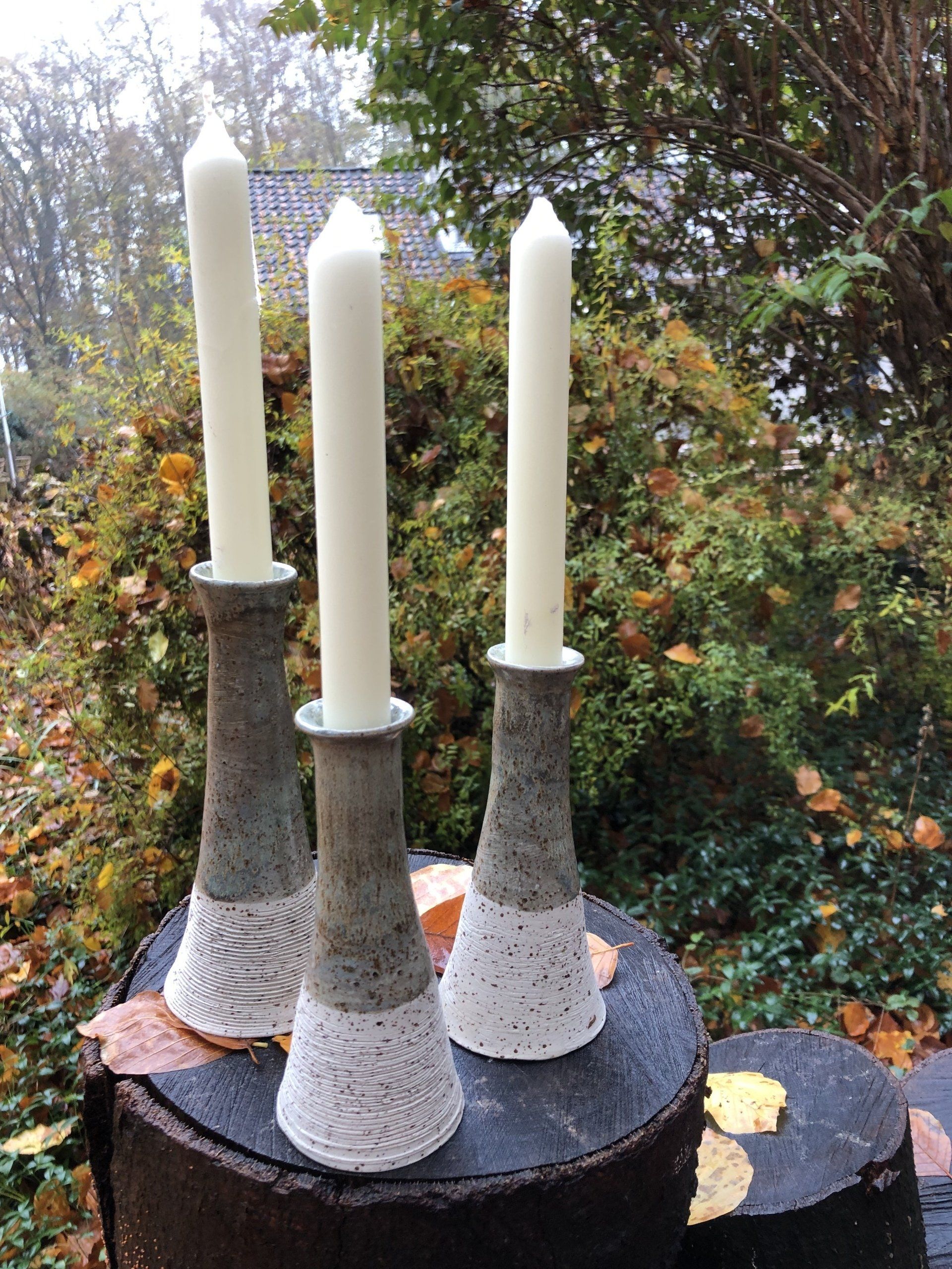 Drie keramische kandelaars met brandende witte kaarsen op een boomstronk voor een groene achtergrond.