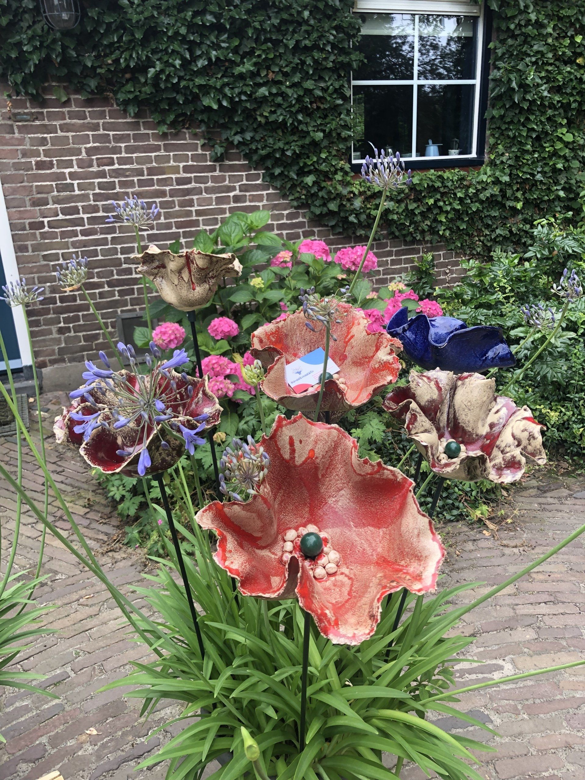 Keramische klaproossculpturen in een tuin, in rode en bruine tinten, met een bakstenen muur en een raam op de achtergrond.