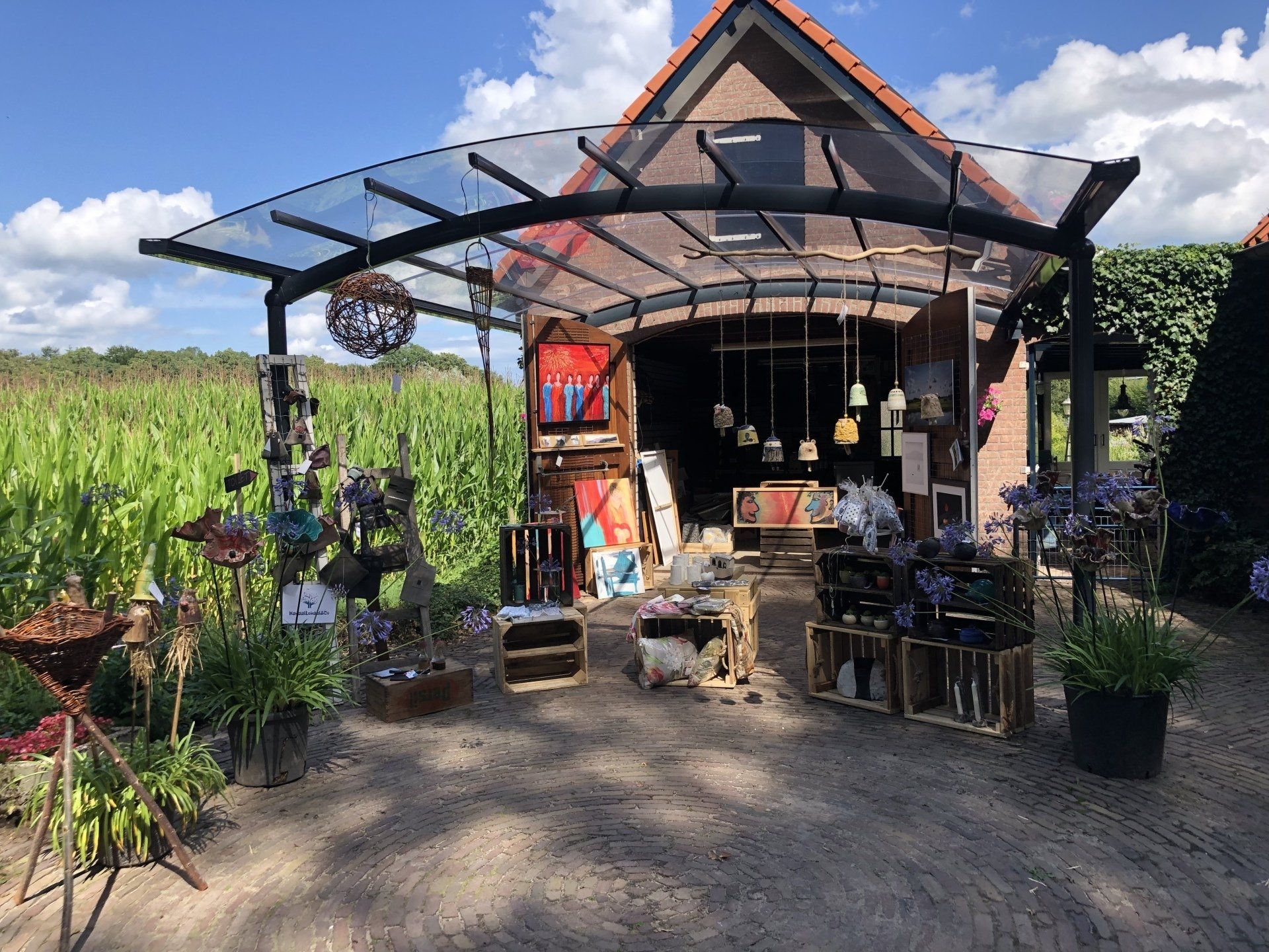 Een openluchtwinkel met een glazen dak waar handgemaakte producten worden tentoongesteld: planten, kunst en verlichting. Gelegen in een veld op een zonnige dag.