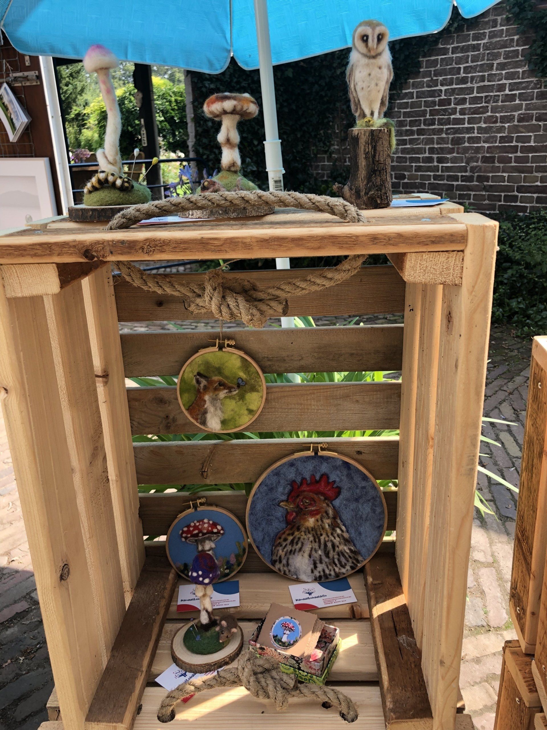 Houten display met kunst: paddenstoelen, een uilenbeeldje, touw en ingelijste dierenborduurwerken.