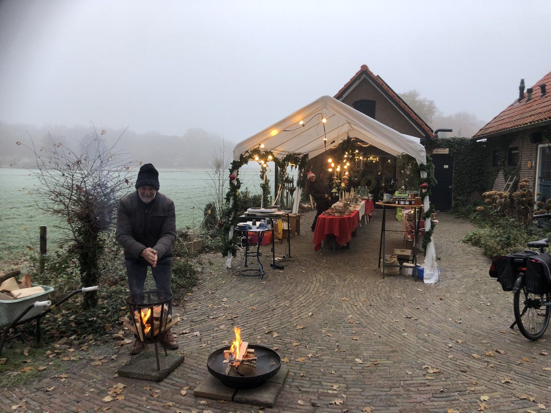 Man bij een vuurplaats met een tafel bedekt met een tent op de achtergrond; een eetgelegenheid in de buitenlucht, mogelijk in de herfst, met een mistig landschap.
