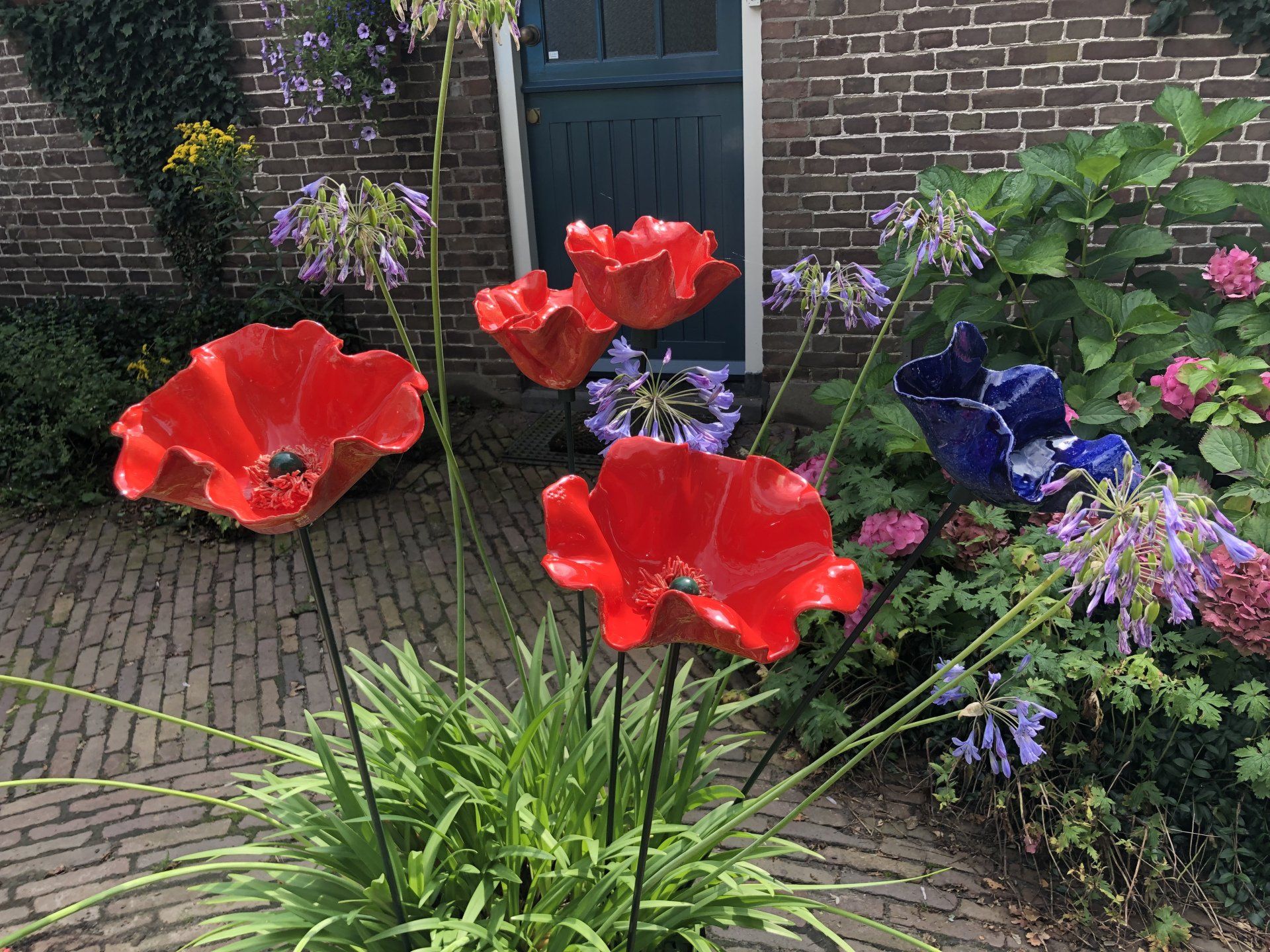 Rode en blauwe keramische bloemsculpturen in een tuin, tegen een bakstenen gebouw en een groene deur.