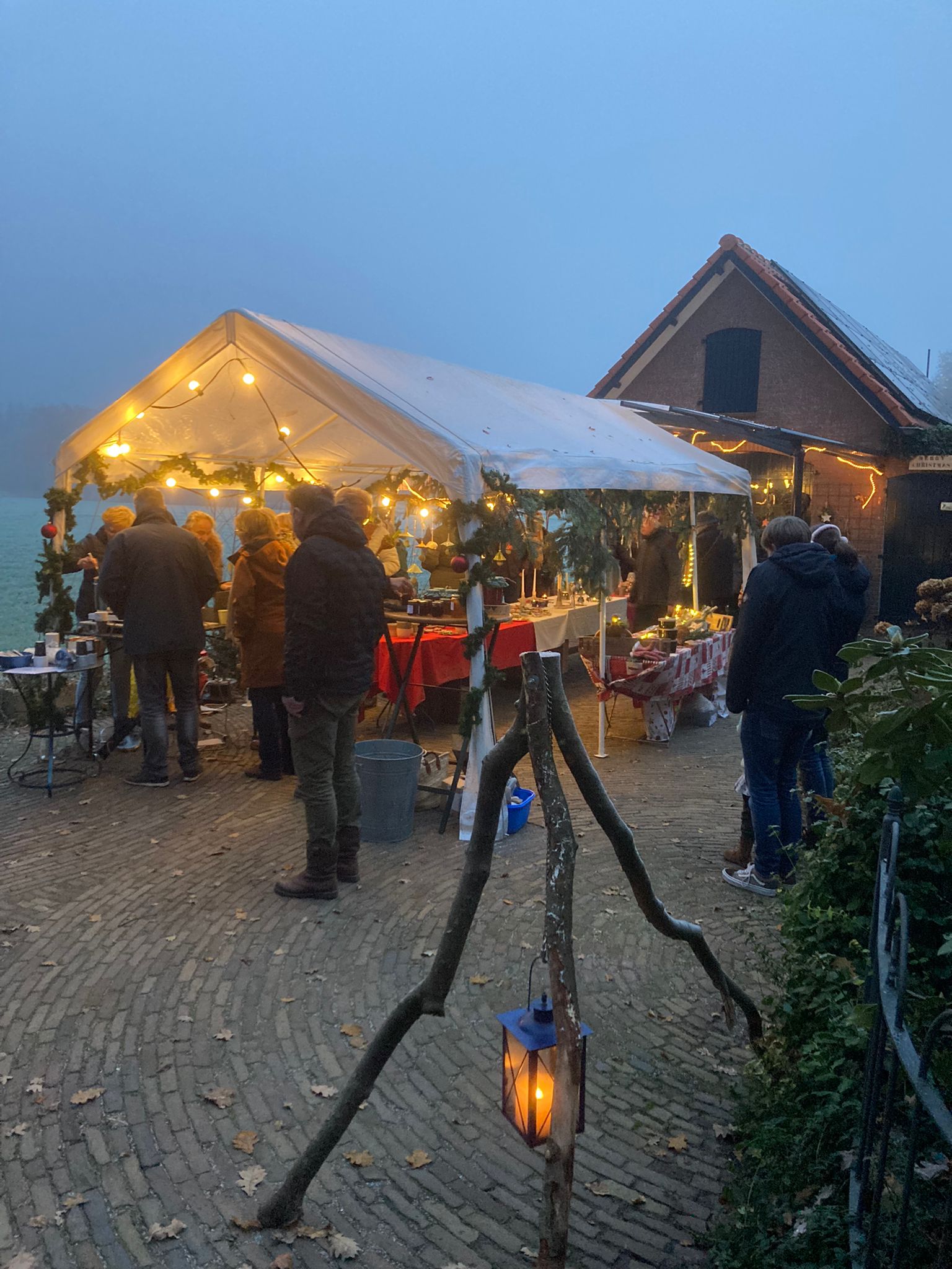 Een openluchtmarkt onder een tent, verlicht met lampen. Mensen kijken rond en een lantaarn hangt op een statief.