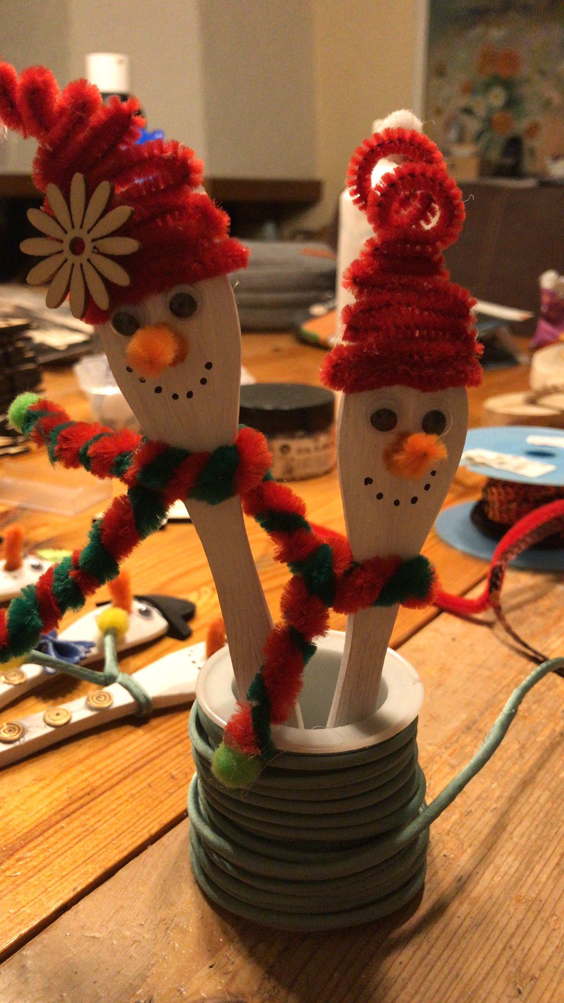 Twee sneeuwpoppen van houten lepels met rode hoedjes en sjaaltjes van pijpenragers, in een houder voor pijpenragers.