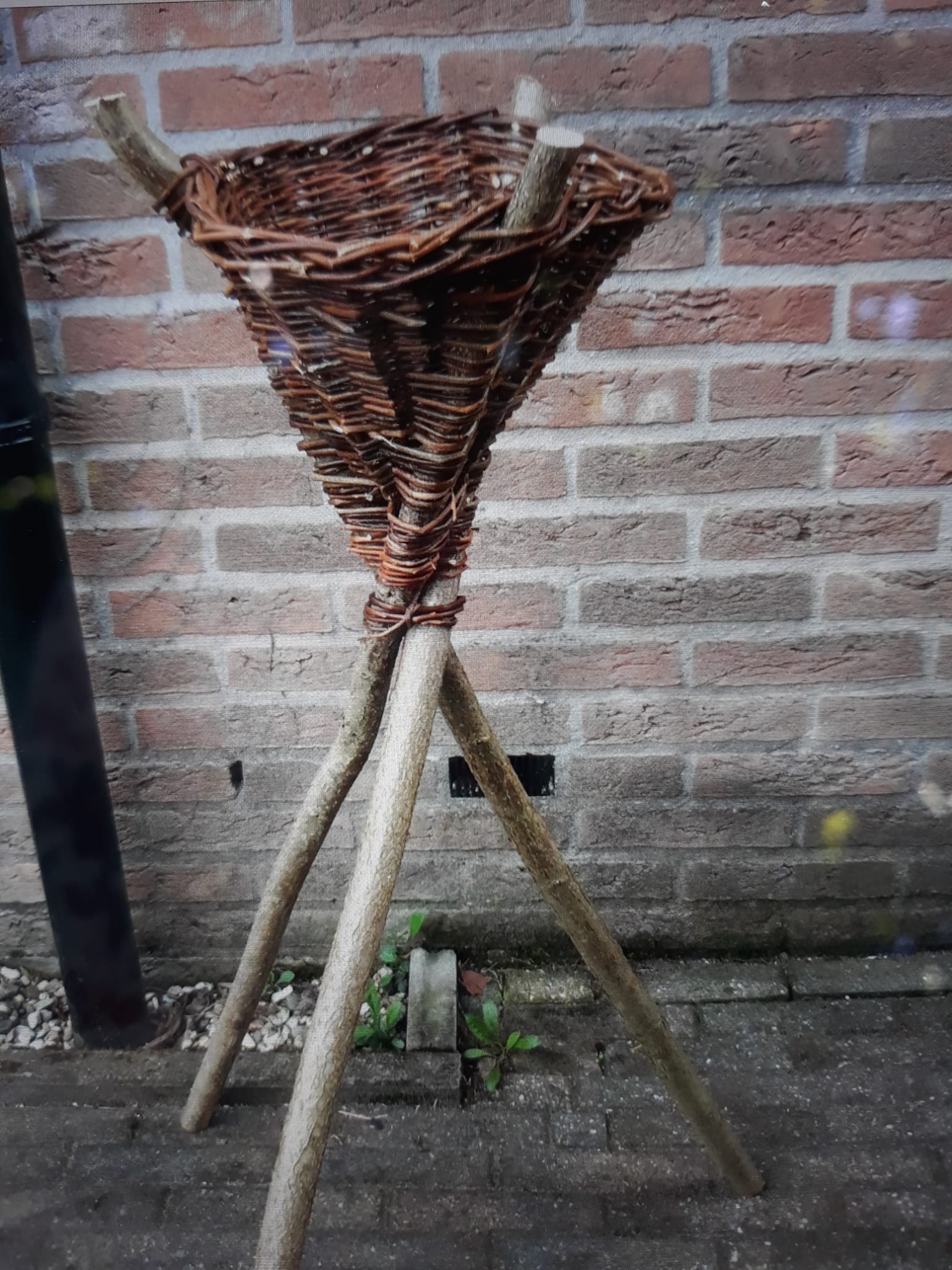Een rieten mand op drie houten poten, geplaatst tegen een bakstenen muur.