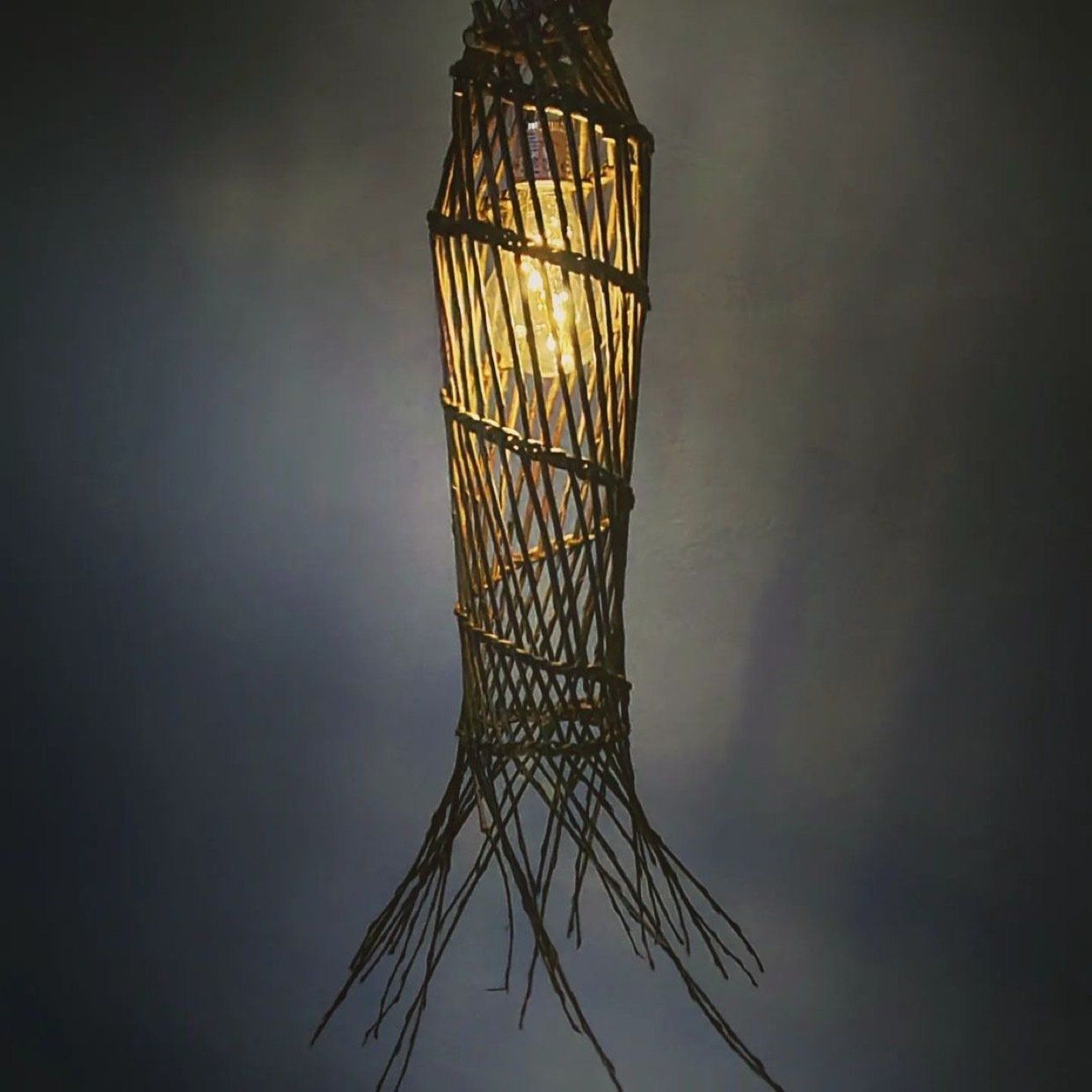 Hanglamp met gevlochten takken en zichtbare lamp, die licht werpt op een grijze muur.
