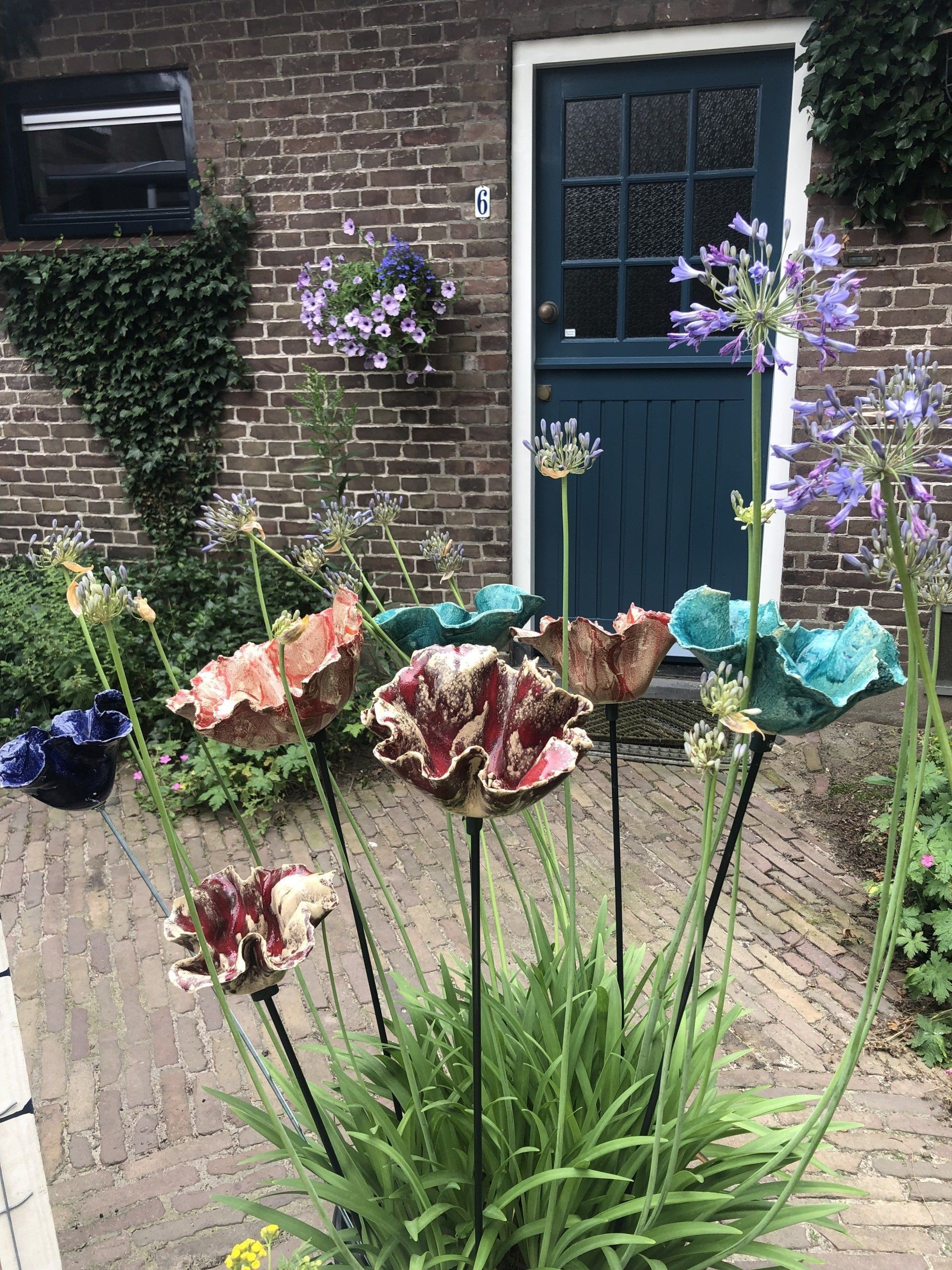 Kleurrijke keramische bloemsculpturen in een tuin, met een bakstenen muur en een turquoise deur op de achtergrond.