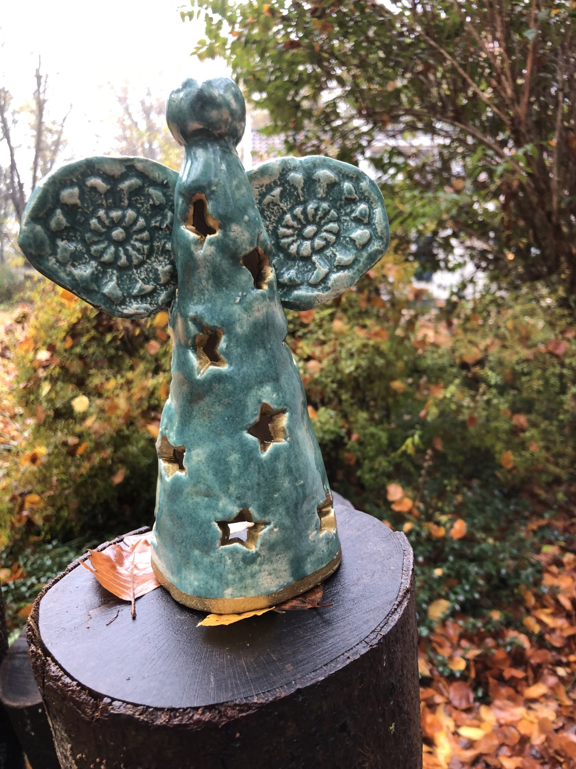 Keramische engel met een turquoise glazuurlaag en stervormige uitsparingen, zittend op een donkere paal, te midden van bladeren.