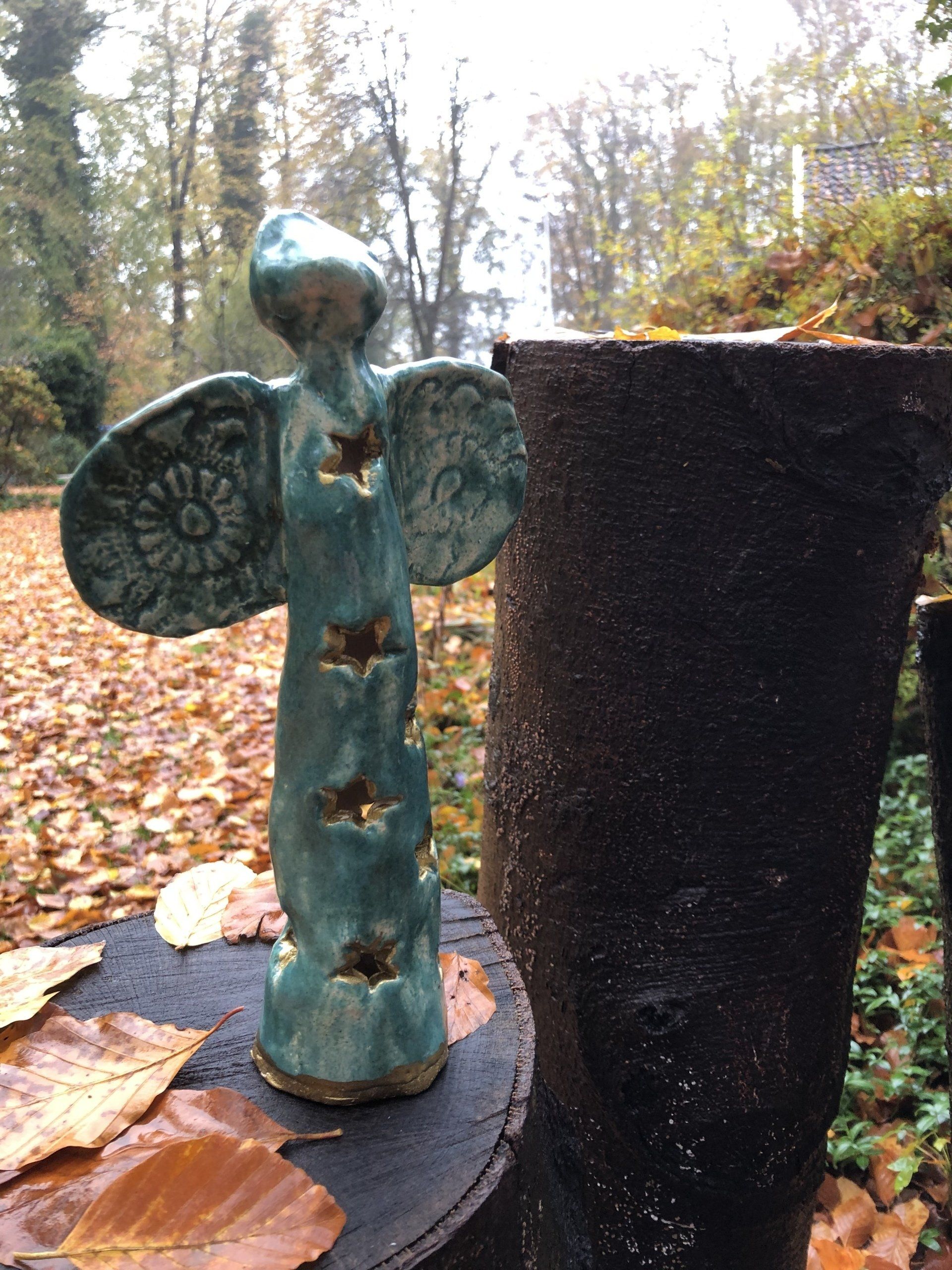 Een turquoisekleurig keramisch engeltje met stervormige uitsparingen, dat in de herfst buiten staat naast een donker, gestructureerd object.