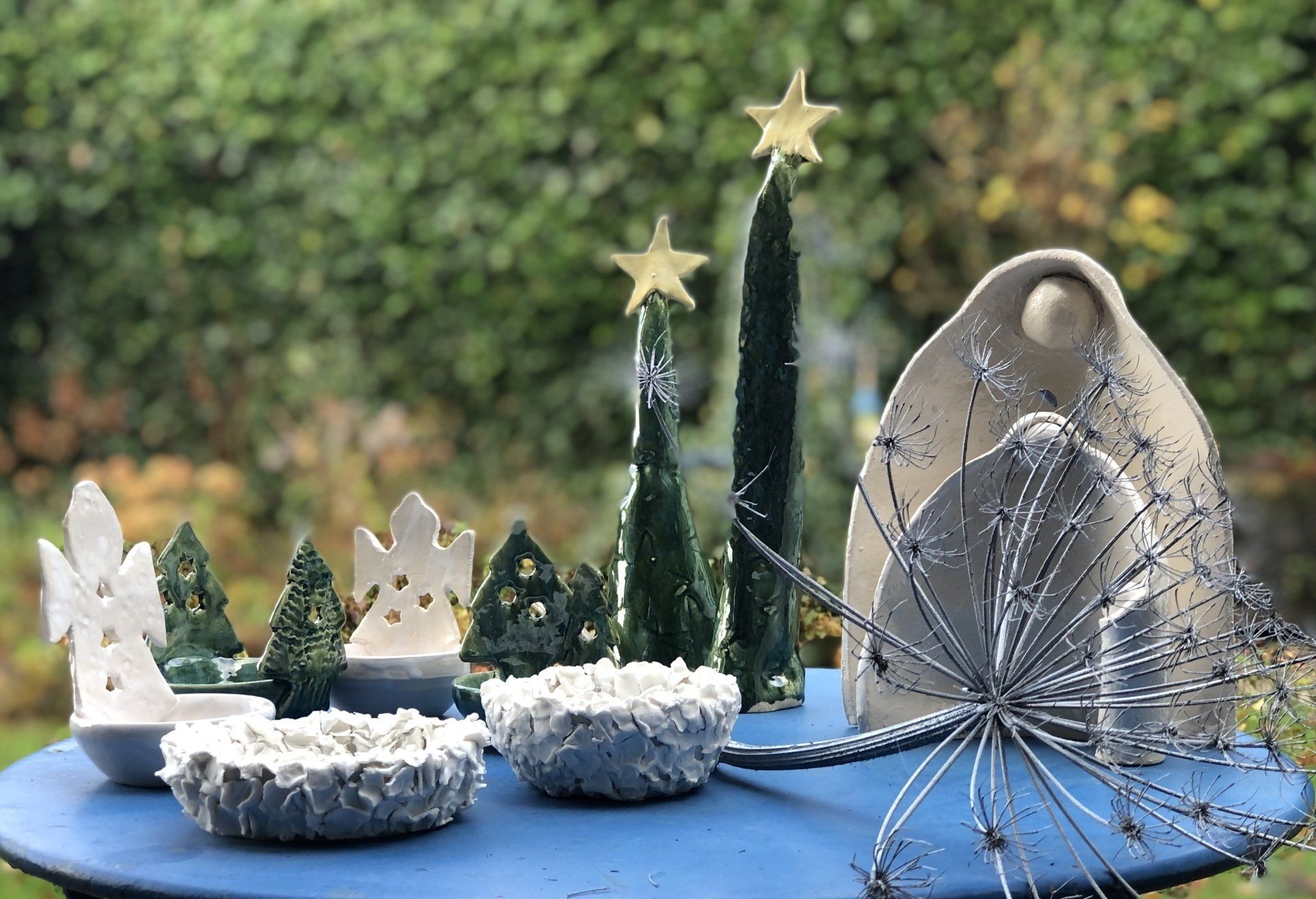 Keramische kerstscène op een blauwe tafel. Met bomen, sterren, engelen en schalen, tegen een natuurlijke achtergrond.
