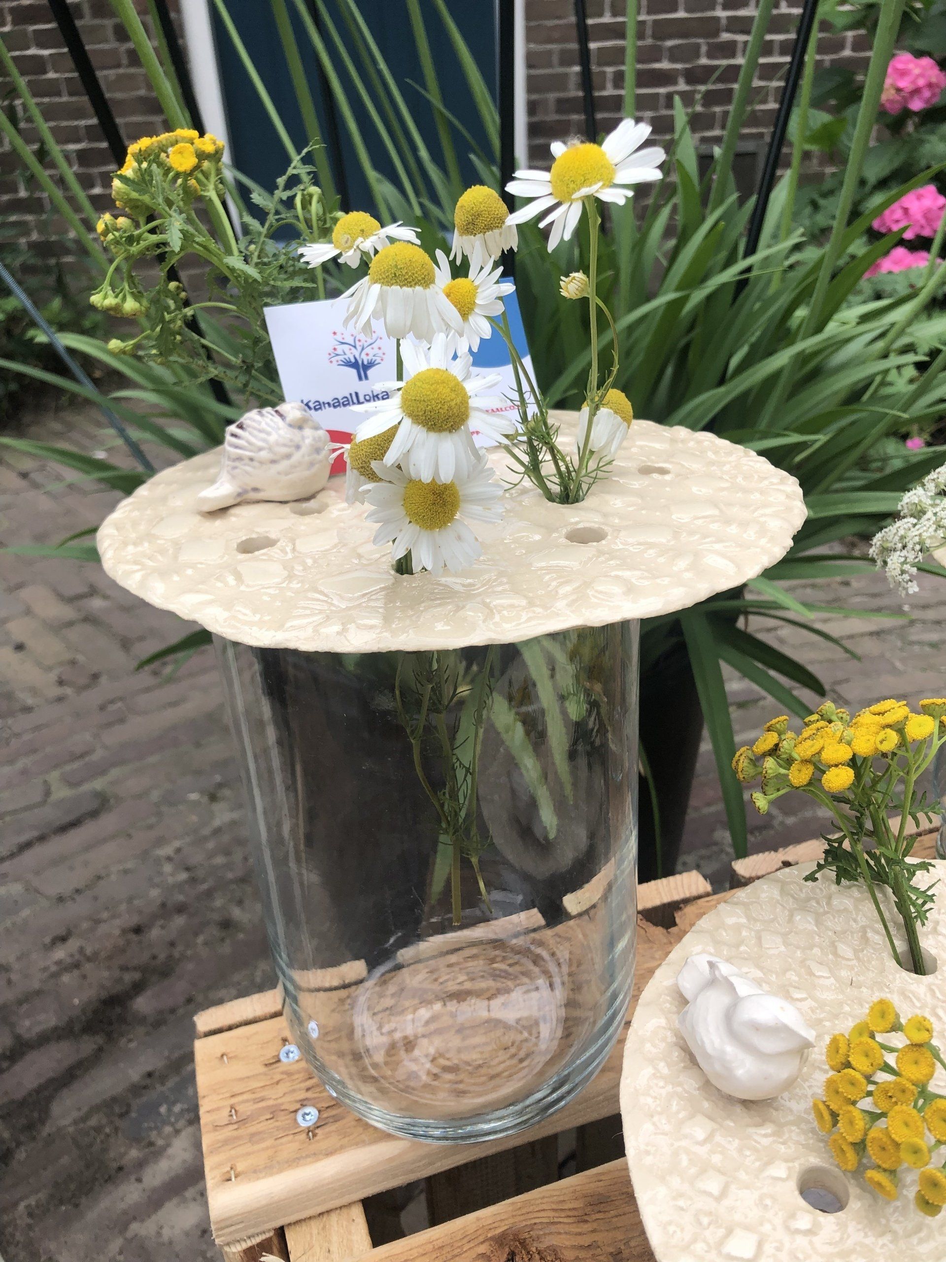 Grote glazen vaas met madeliefjes, met een decoratief deksel in de vorm van een zeeschelp, op een houten pallet buiten.