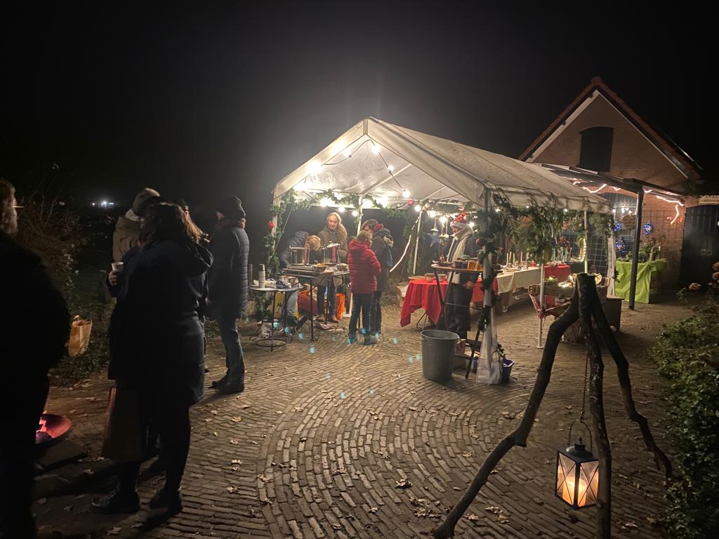 Avondmarkt met tent, verlichting, mensen en een gebouw op de achtergrond. Vuurplaats met lantaarn op de voorgrond.