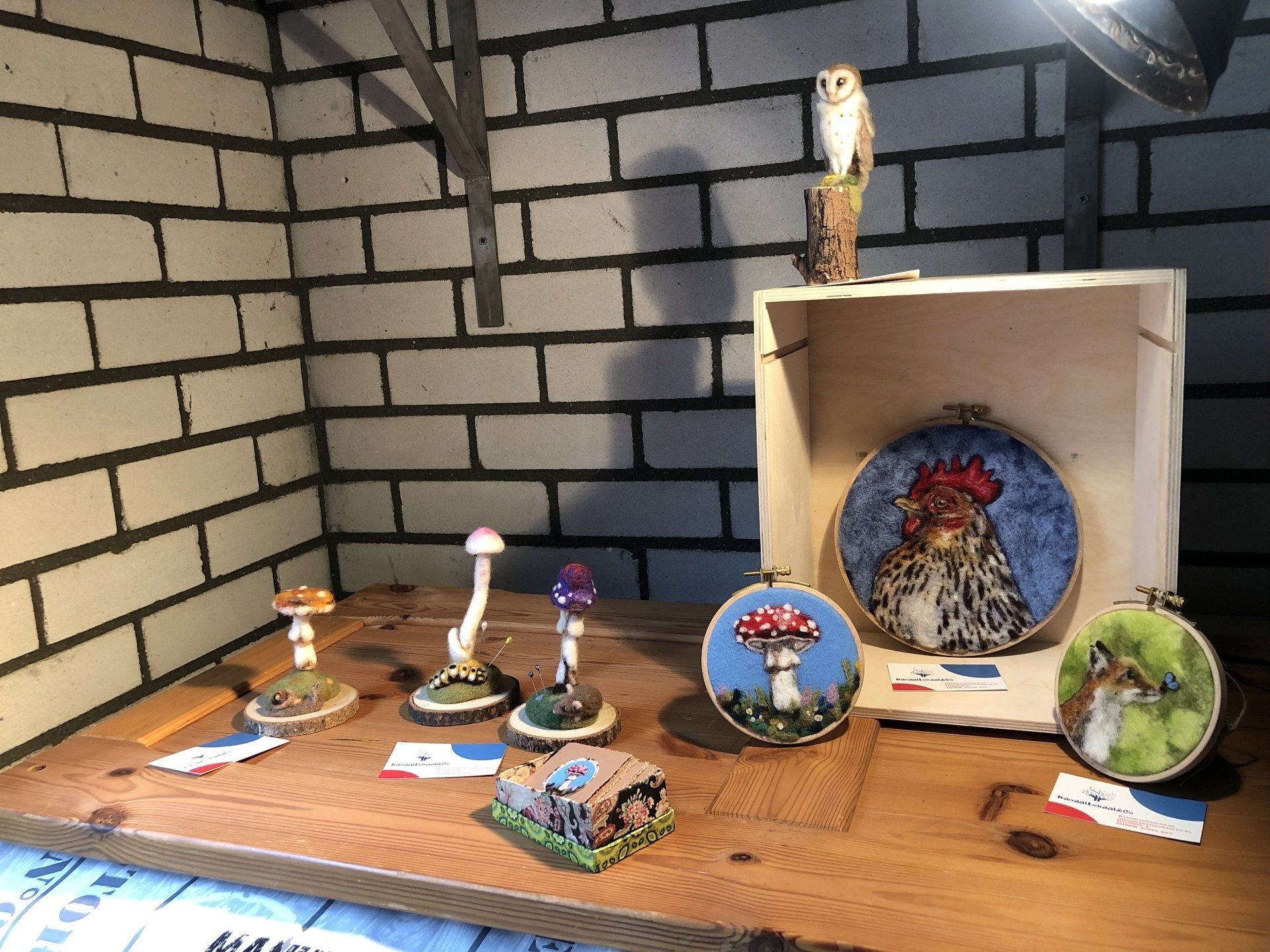 Tentoonstelling van handgemaakte kunstwerken: borduurringen, paddenstoelsculpturen, een houten uil en een achtergrond met een bakstenen muur.