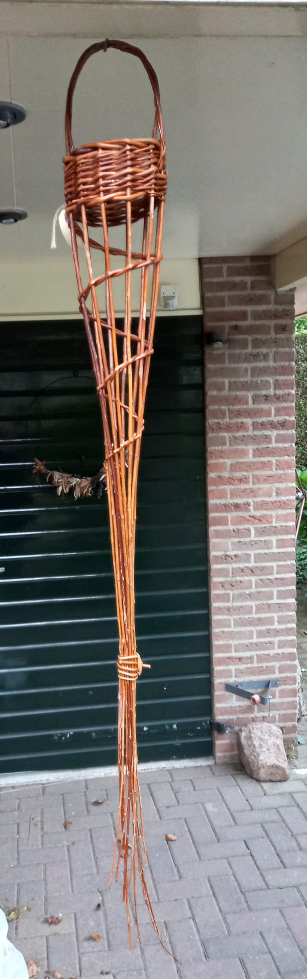 Een gevlochten mand hangt onder een veranda. De mand loopt taps toe van een ronde bovenkant naar een punt. Op de achtergrond zijn een bakstenen muur en een groene garagedeur te zien.