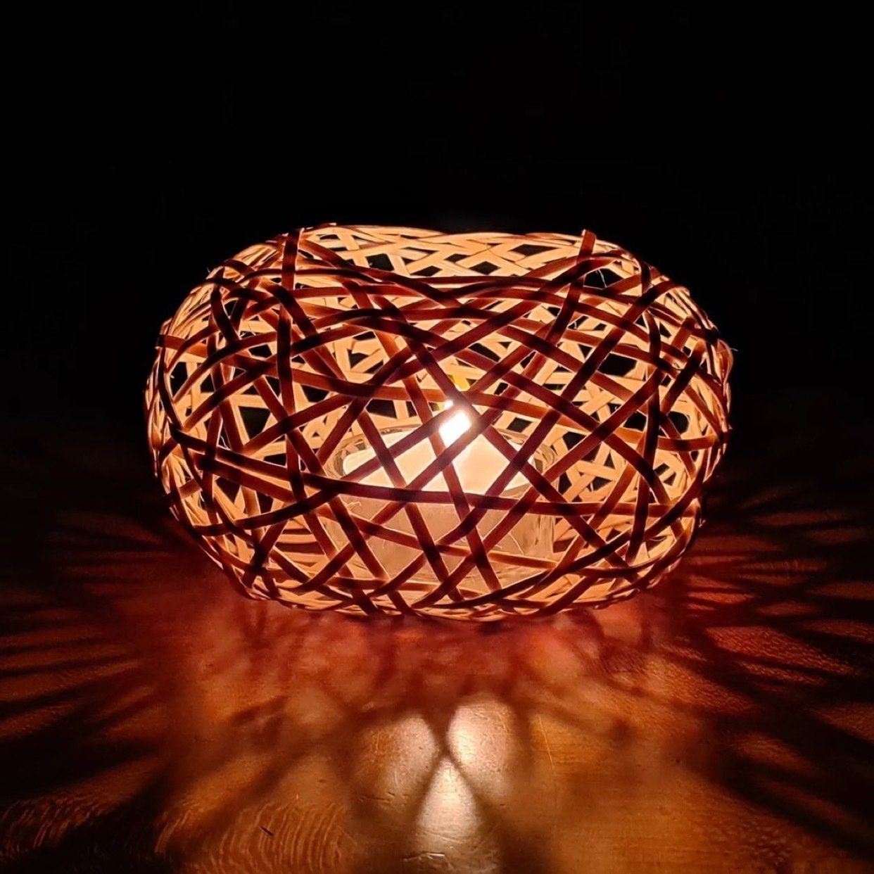 Een kaarsenhouder van geweven bamboe met een brandende kaars erin, die een warm lichtspel en schaduwen verspreidt.
