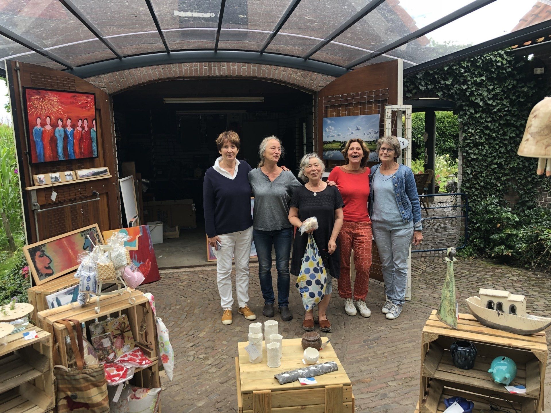 Vijf vrouwen staan voor een open garage waar kunstwerken tentoongesteld staan. In kratten liggen aardewerk en schilderijen; de lucht is bewolkt.