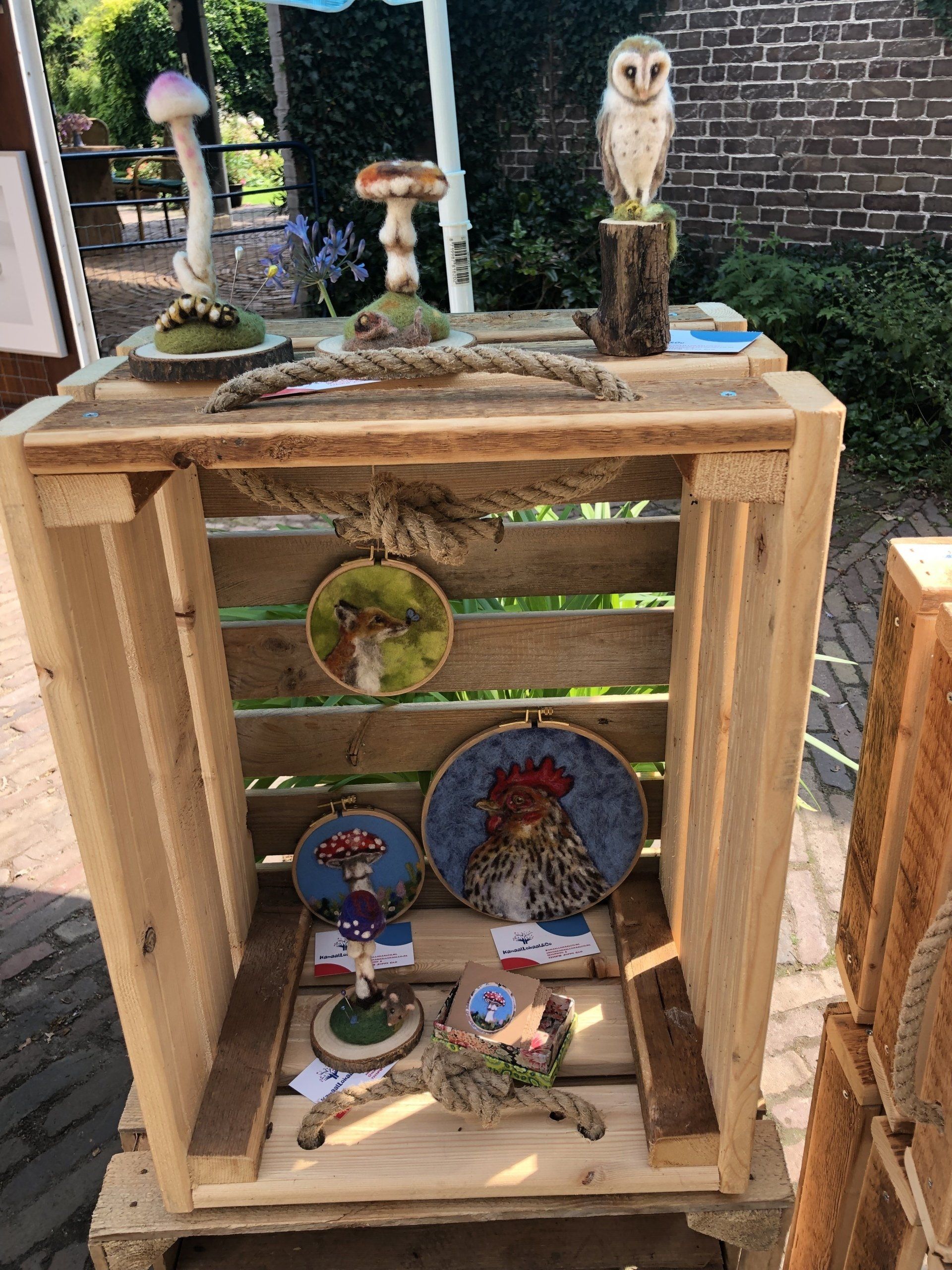 Een houten kist waarin handgemaakte kunstwerken van paddenstoelen en uilen worden tentoongesteld.
