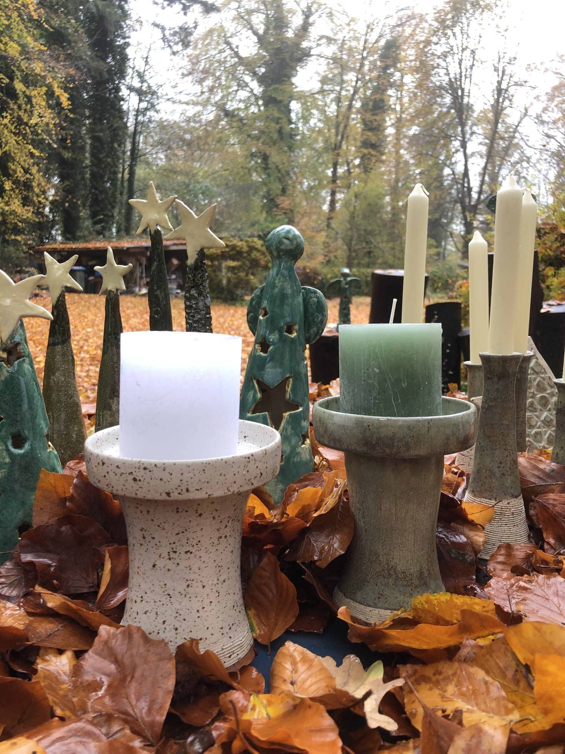 Kaarsen in keramische houders tussen gevallen bladeren, met abstracte sculpturen in de buitenlucht op de achtergrond.