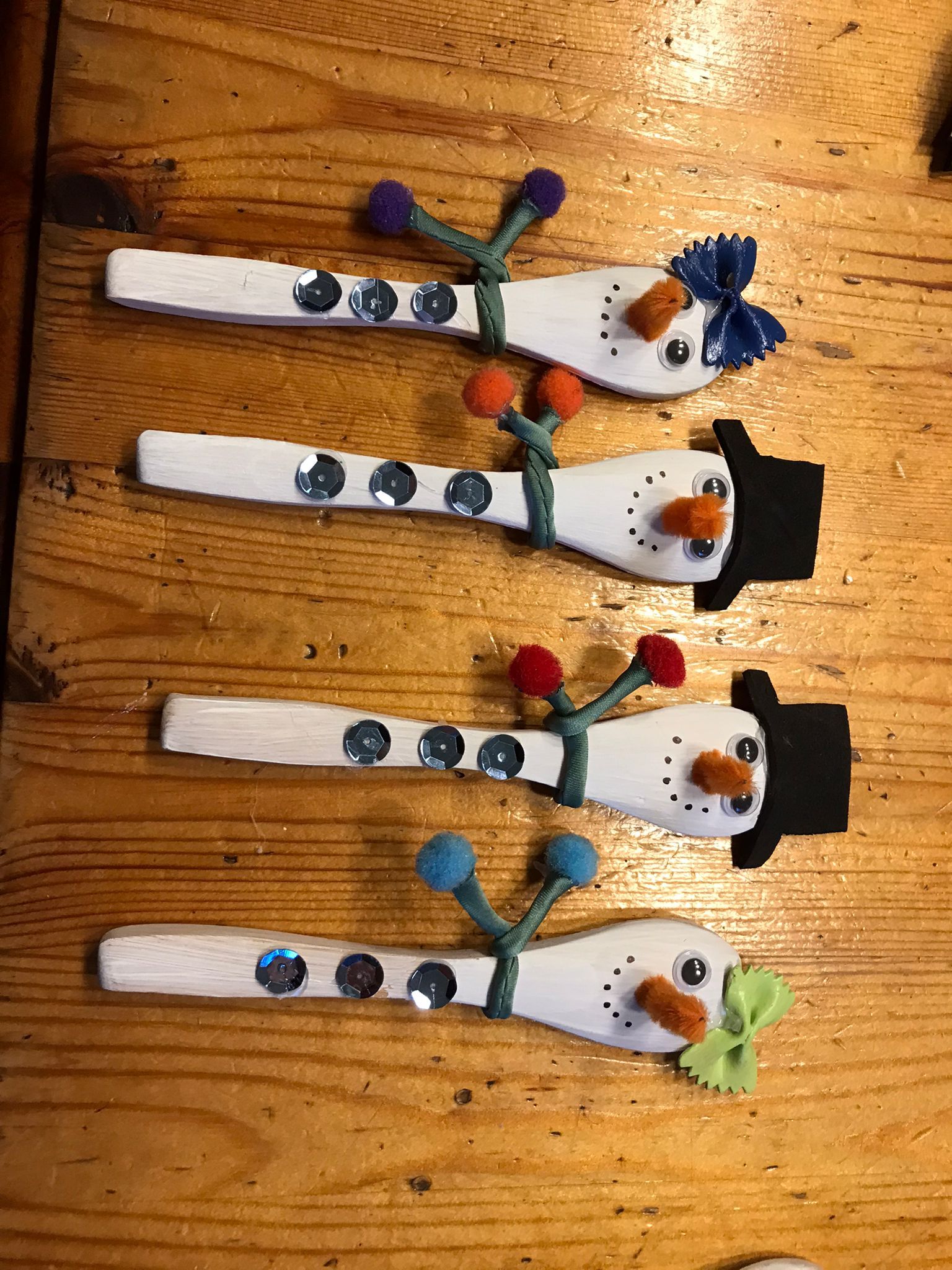 Op een houten tafel staan vier sneeuwpoppen, gemaakt van lepels, elk met een andere versiering.