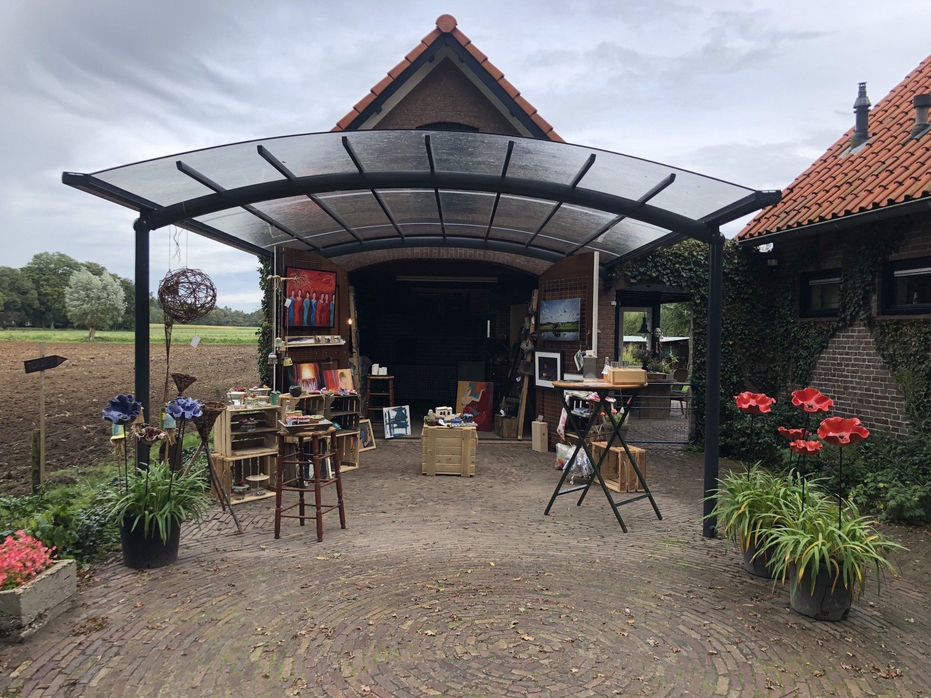 Kunsttentoonstelling onder een afdak, met schilderijen, aardewerk en bloemen buiten een bakstenen gebouw.