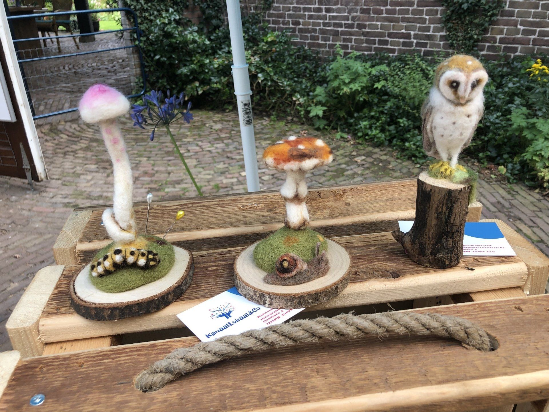 Drie kunstwerken van wol: een paddenstoel en een uil op houten sokkels, tentoongesteld op een houten tafel.